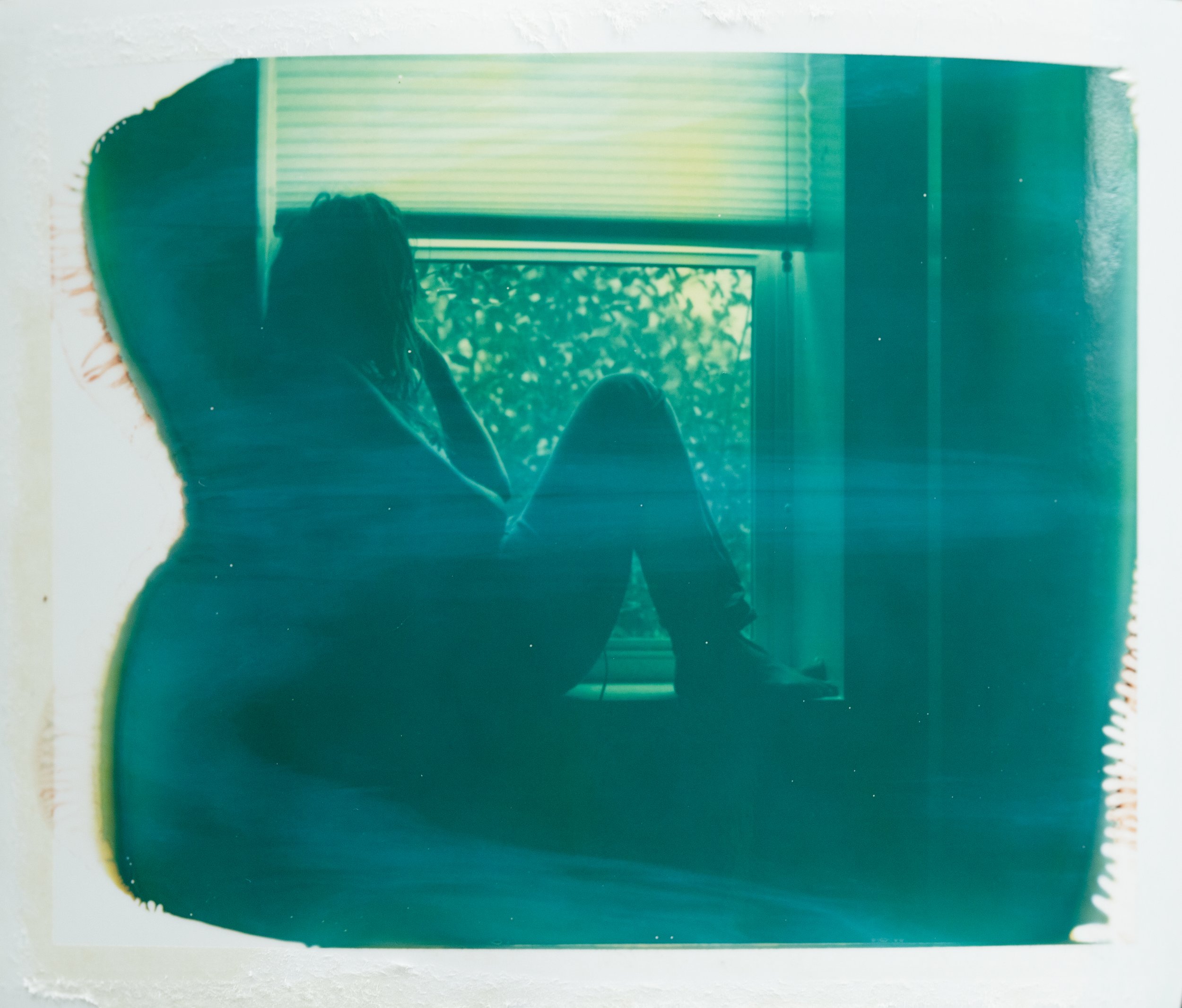 Silhouette of a person sitting on a windowsill, looking outside, with blinds partially open, revealing trees outside.