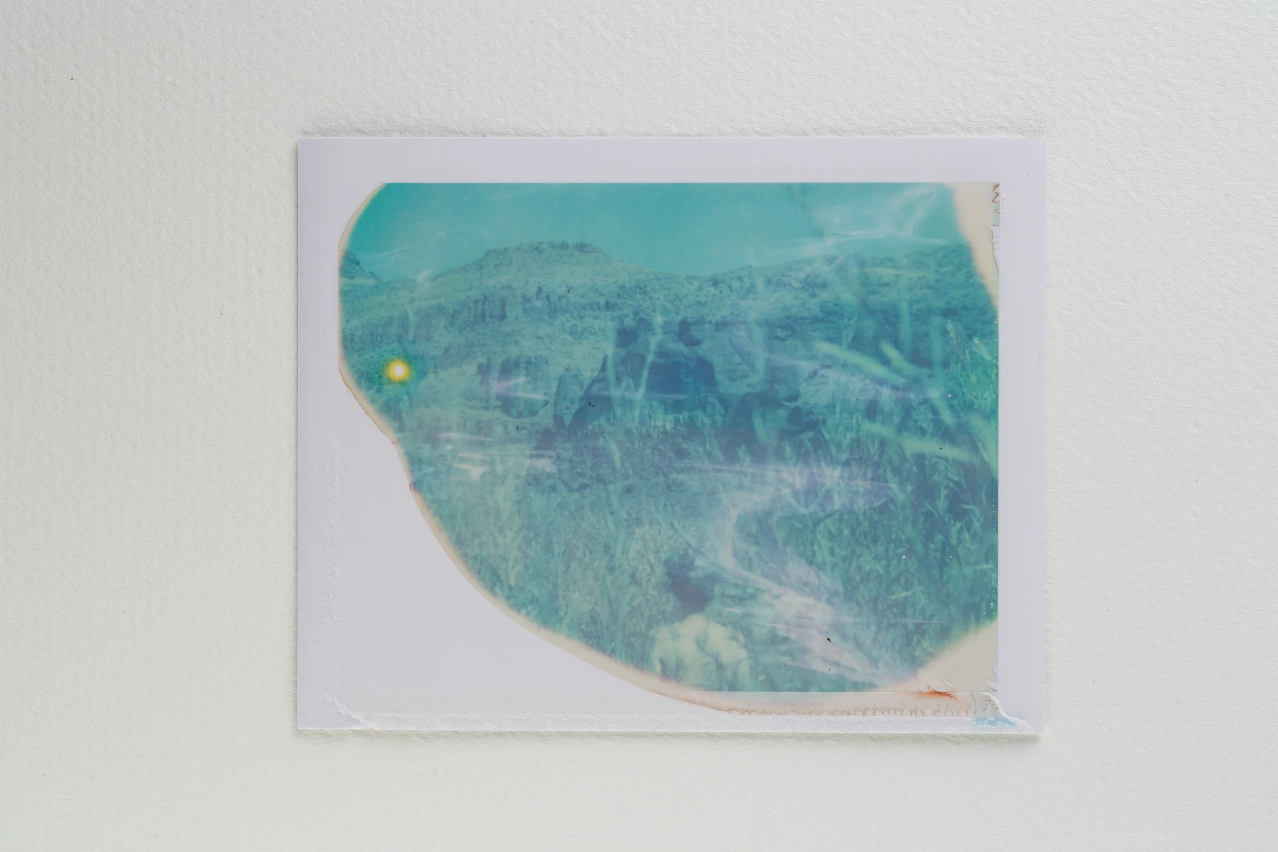 A damaged, color-tinted photo of a mountainous landscape with greenery and hills, mounted on a white wall.