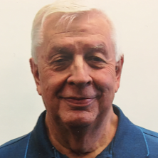 An elderly man with white hair wearing a dark blue collared shirt, smiling slightly, against a plain light background.