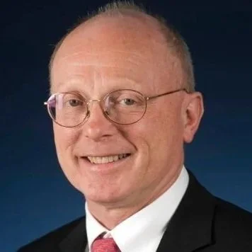 A middle-aged man with glasses, short light hair, wearing a black suit, white shirt, and pink tie, smiling against a dark background.