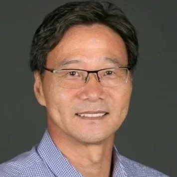 Portrait of a middle-aged man with glasses, wearing a blue checkered shirt, smiling against a gray background.