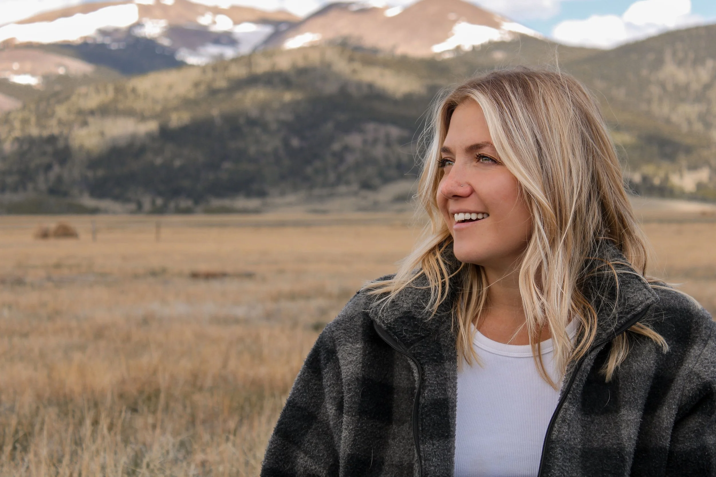 A young woman with wavy blonde hair and a nose piercing smiling outdoors in a field with mountains in the background.