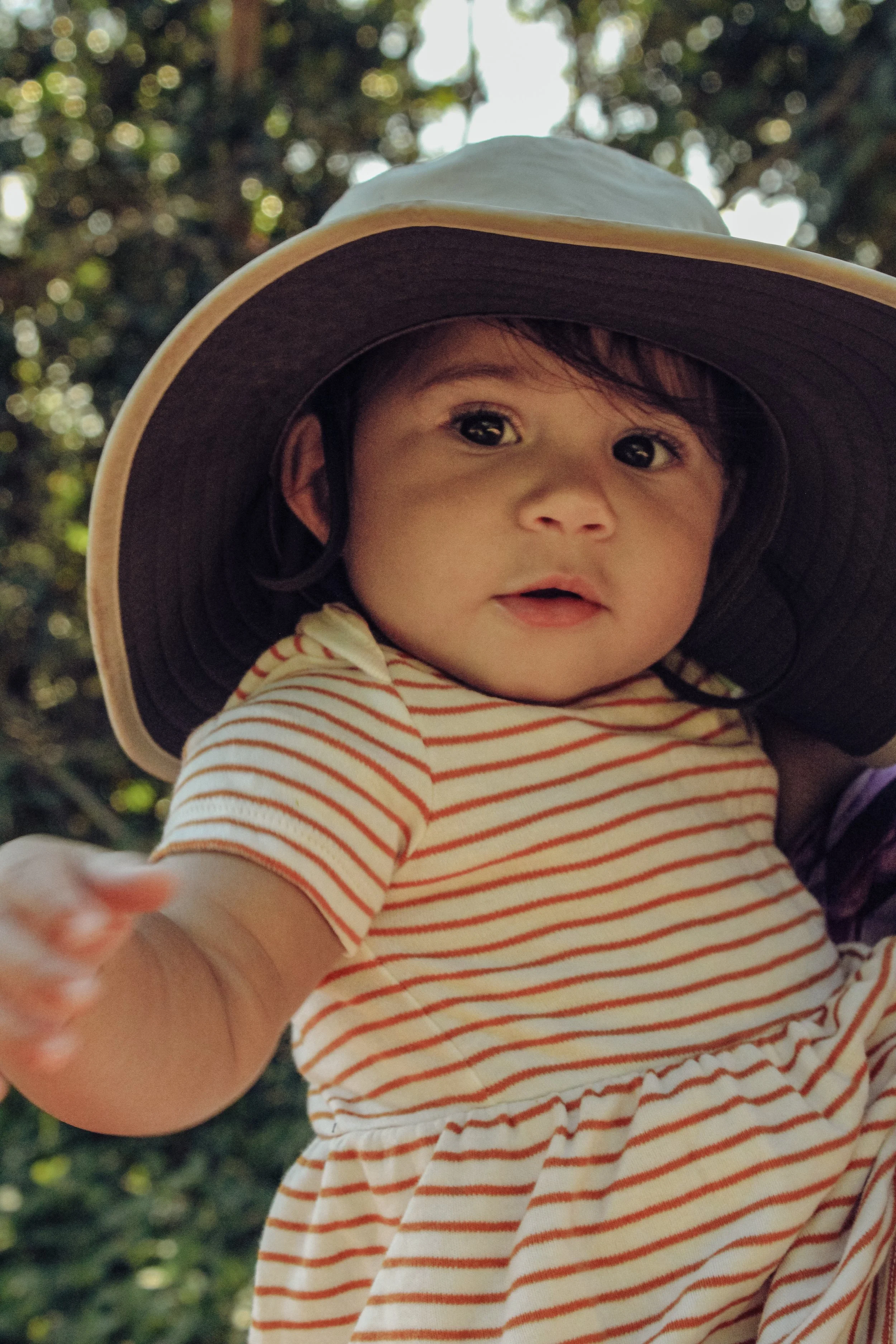 Child with dark hair wearing a striped dress and a large sun hat, reaching forward outdoors with blurred greenery in background.