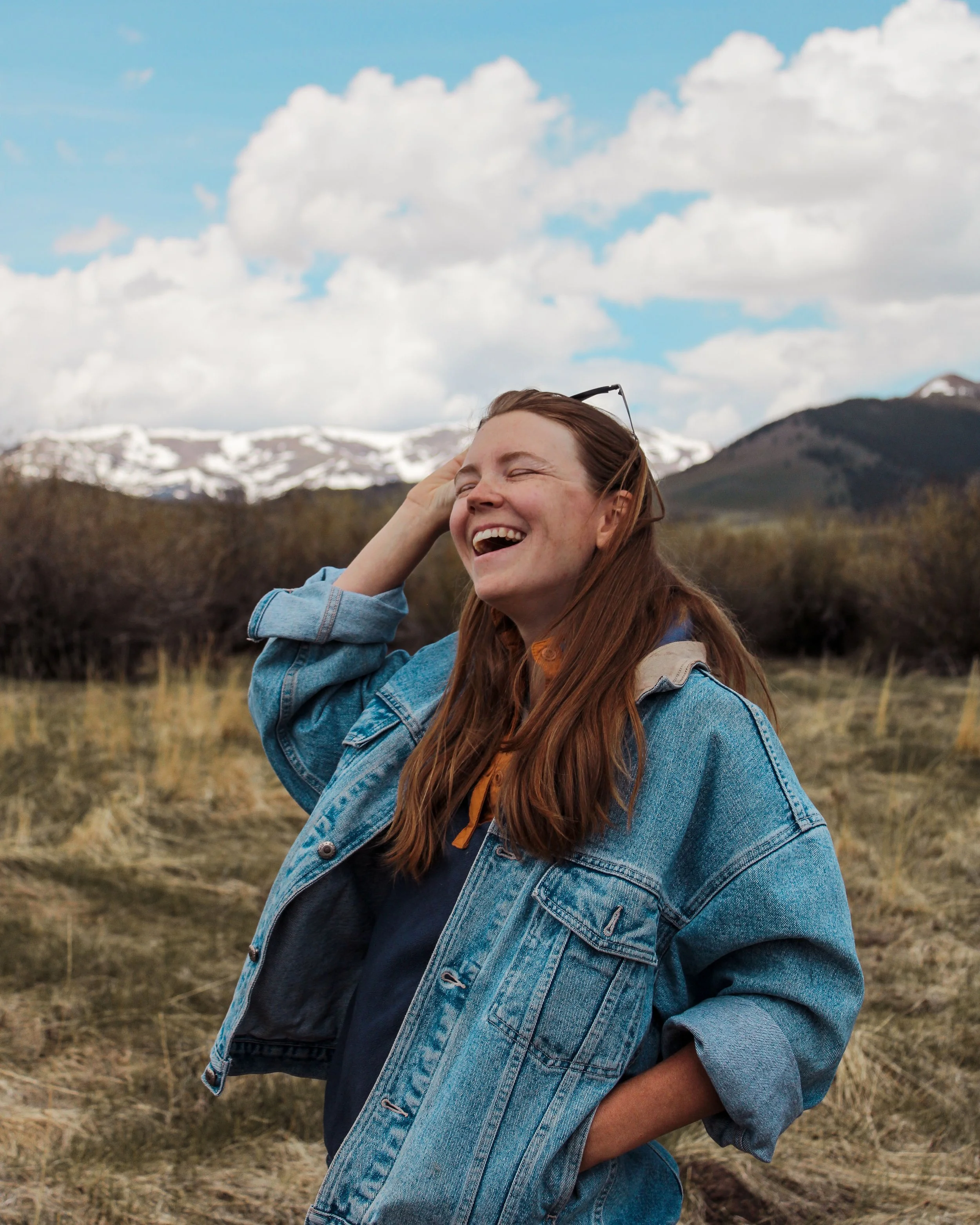 A young woman with long brown hair wearing a denim jacket smiling and laughing outdoors with mountains and a partly cloudy sky in the background.