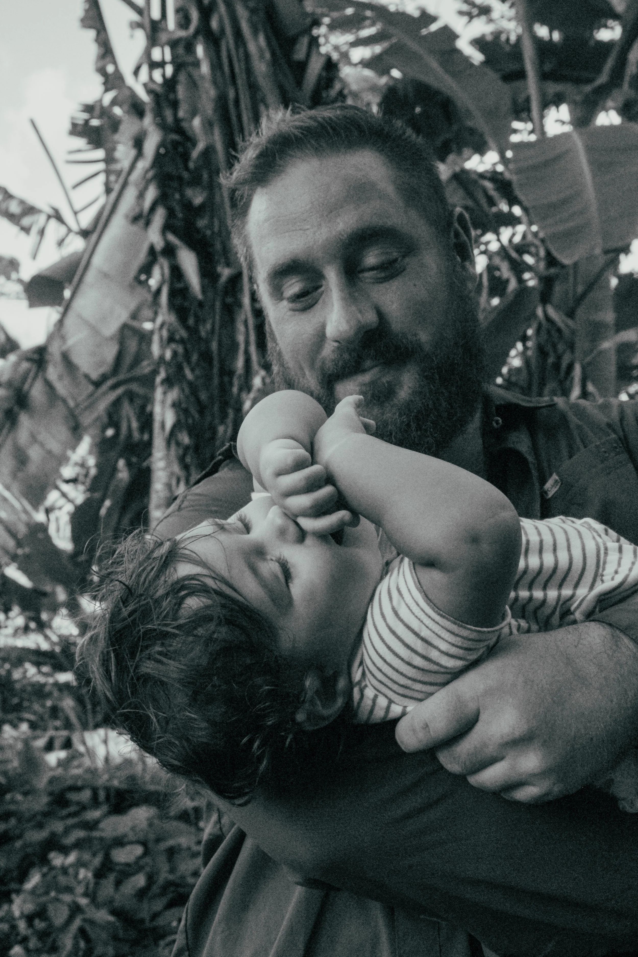 A bearded man holding a sleeping toddler and gently touching the child's face, outdoors with trees in the background.