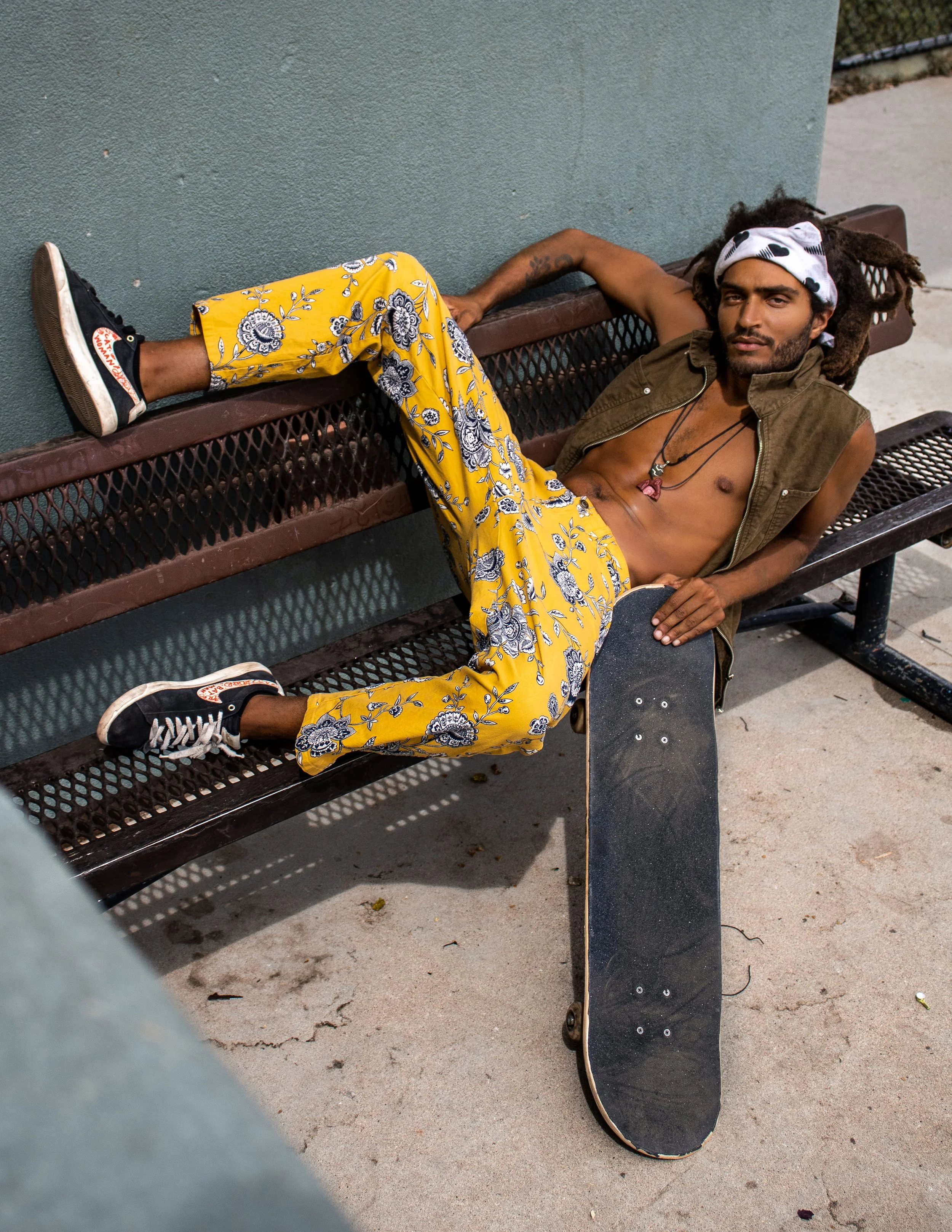 A man with dreadlocks lounging on a park bench, holding a skateboard, wearing yellow floral pants, a sleeveless vest, and sneakers.
