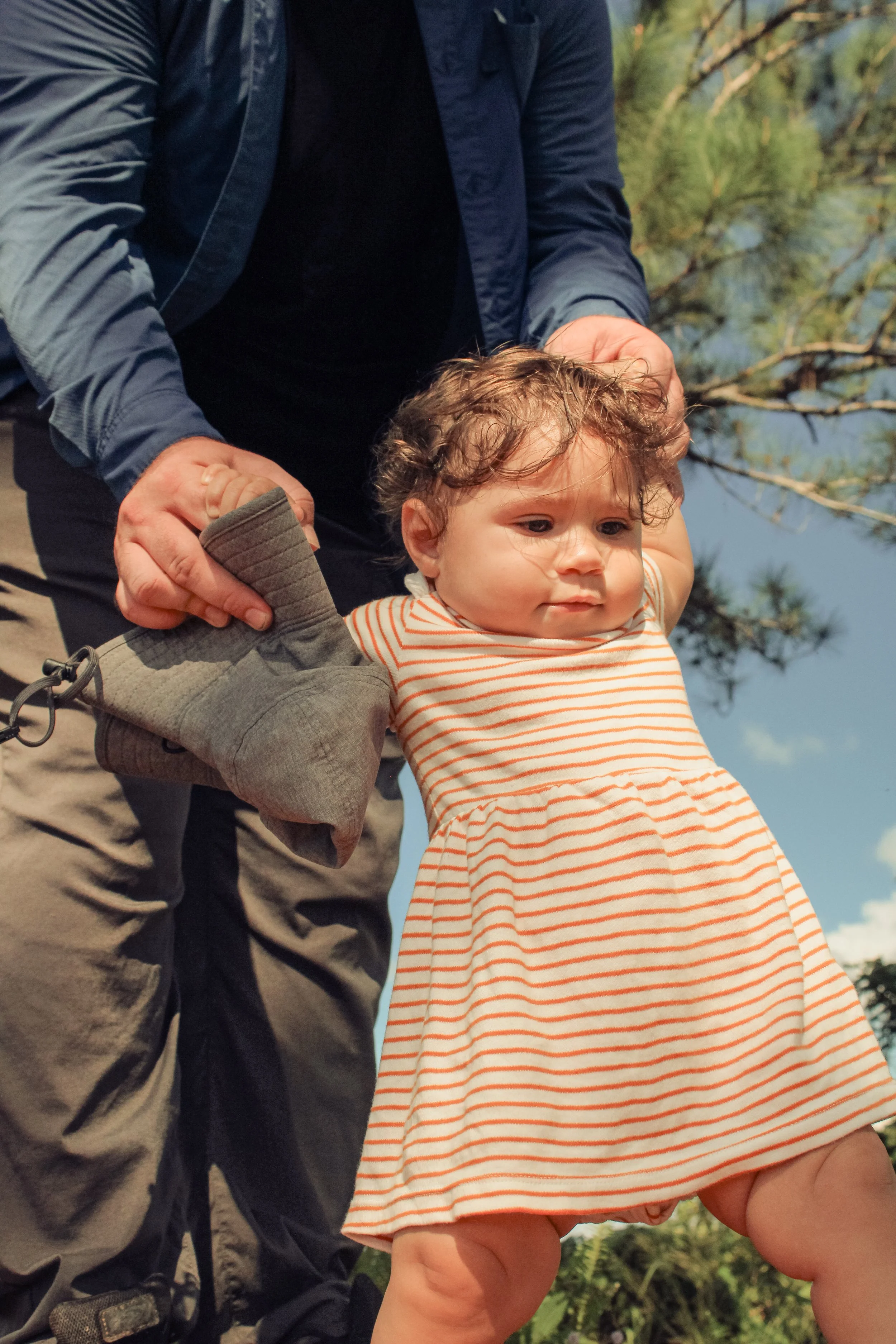A young toddler girl with curly hair, wearing a striped orange and white dress, is being helped by an adult with a sock, outdoors near trees, under a blue sky.