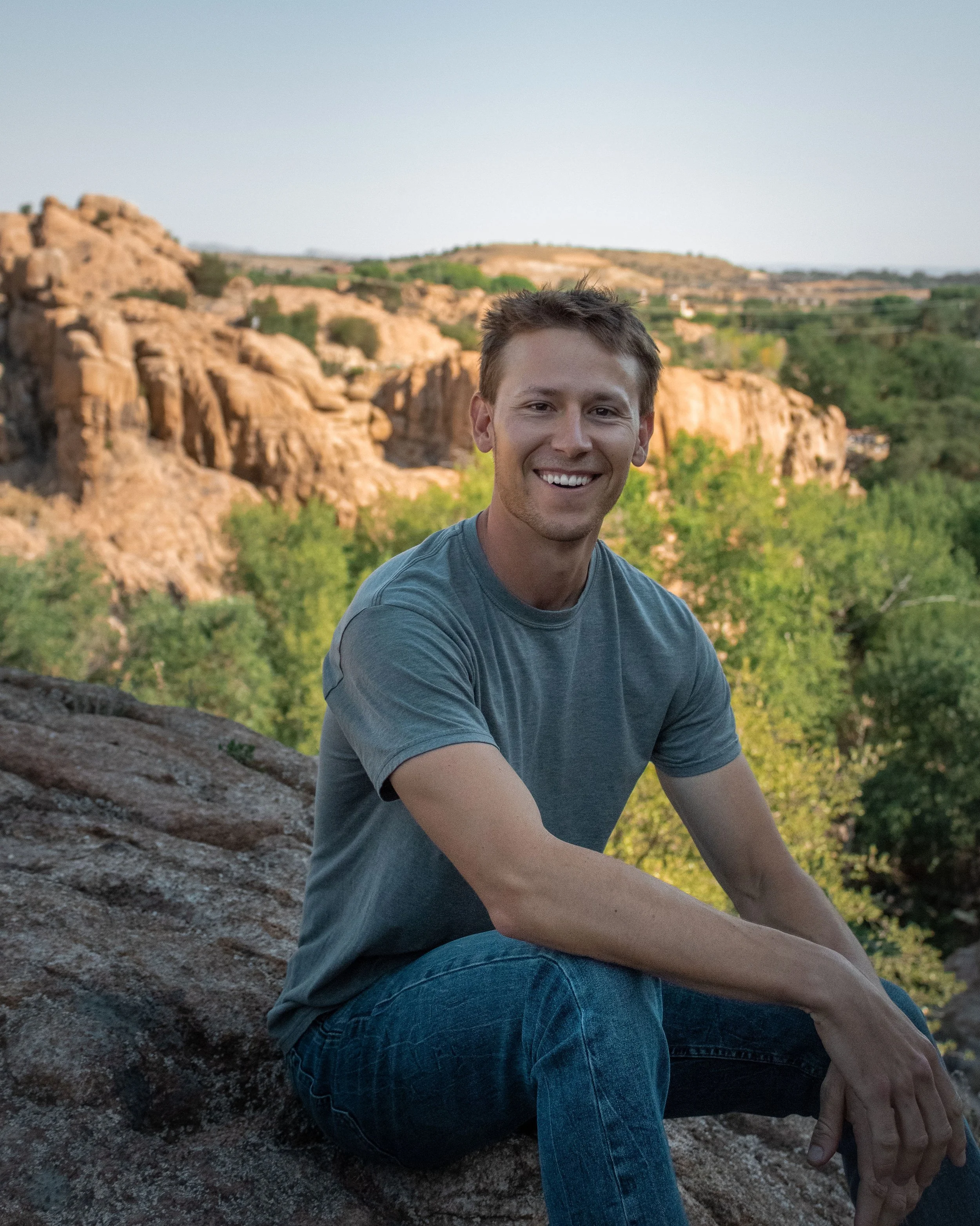 A young man sitting on a rock outdoors with a landscape of rocky hills and trees in the background, smiling at the camera.