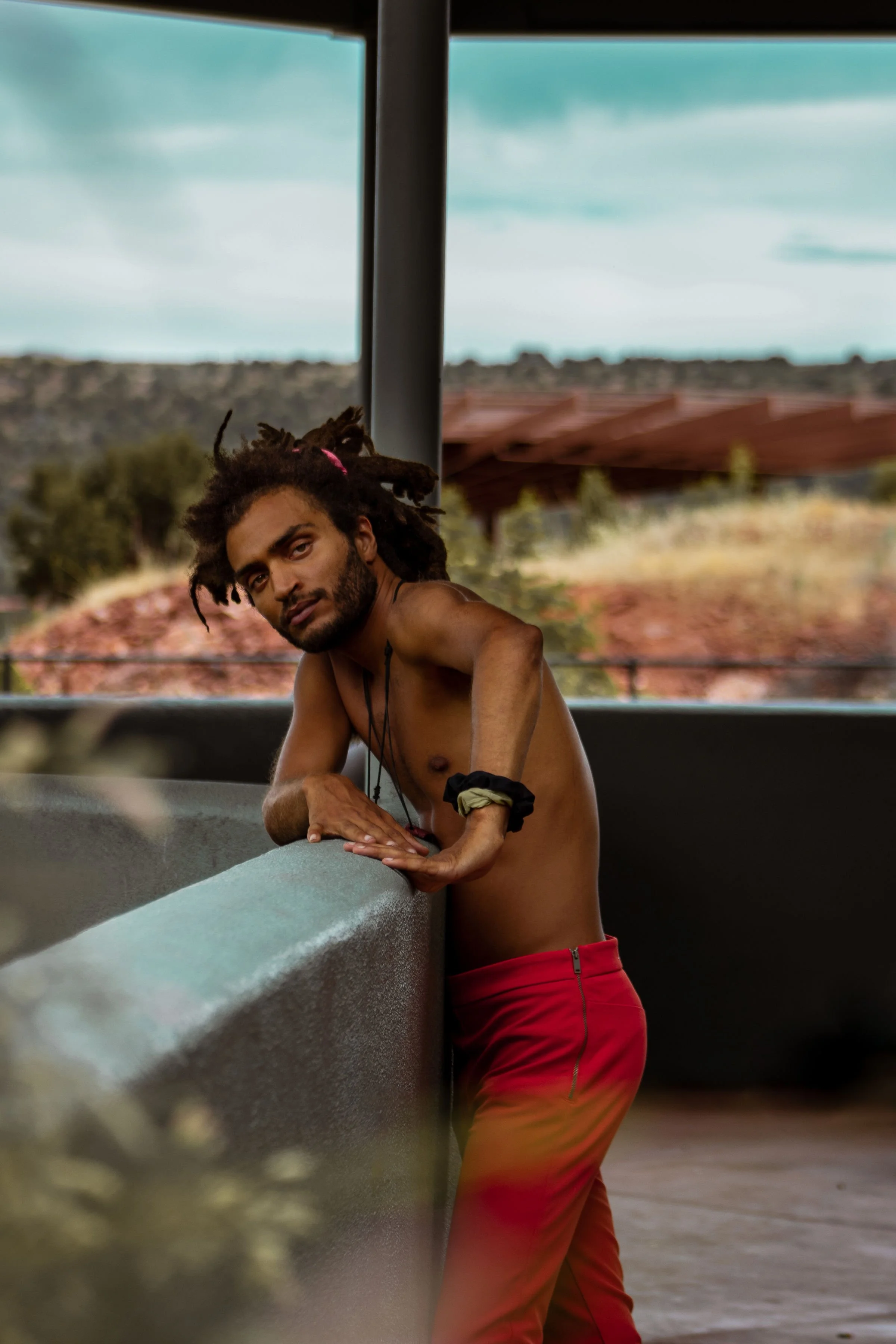 A shirtless man with dreadlocks leaning on a concrete ledge, outdoors with a blurred background of landscape and wooden structures.