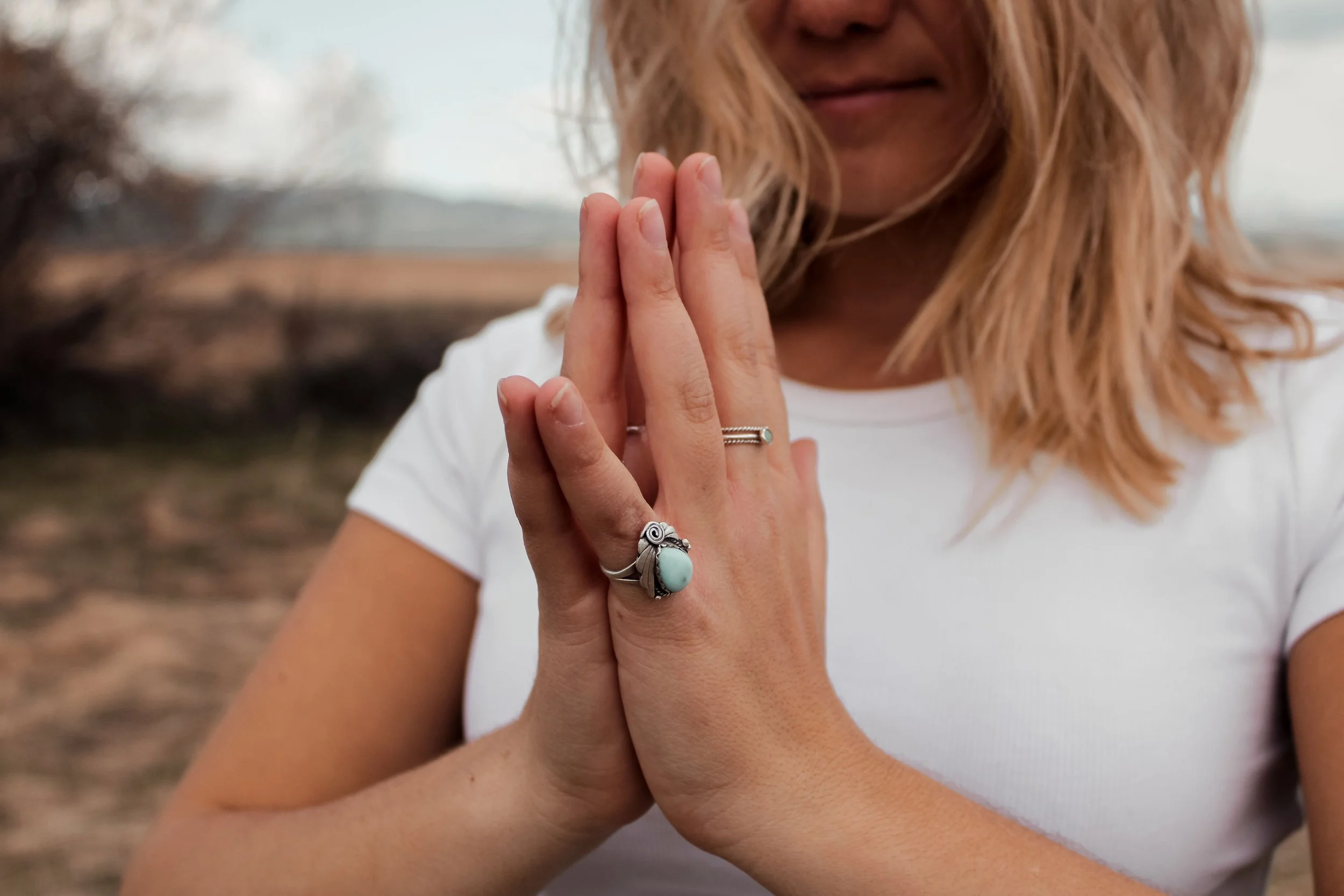 A woman with blonde hair in a white shirt positions her hands together in a prayer gesture, showcasing rings on her fingers including a turquoise and silver ring, outdoors with a blurred natural background.