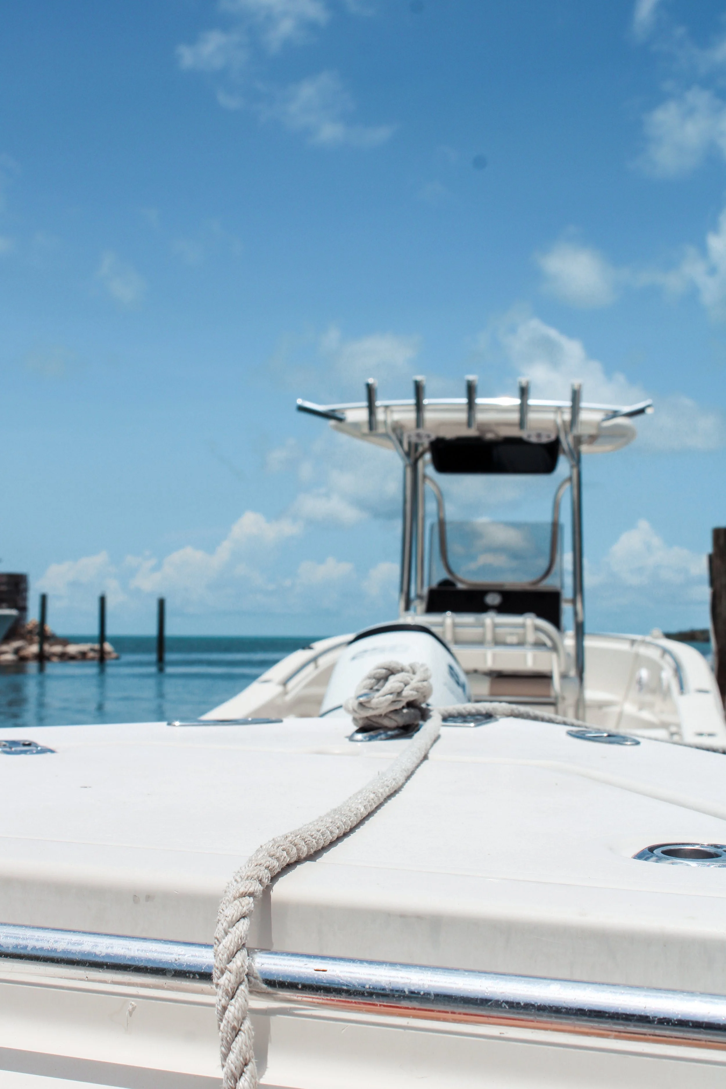 View of a boat docked at a marina with a clear blue sky and some clouds in the background. The boat's bow with a rope tied to its front is visible in the foreground, and the boat's steering console is visible in the middle ground.