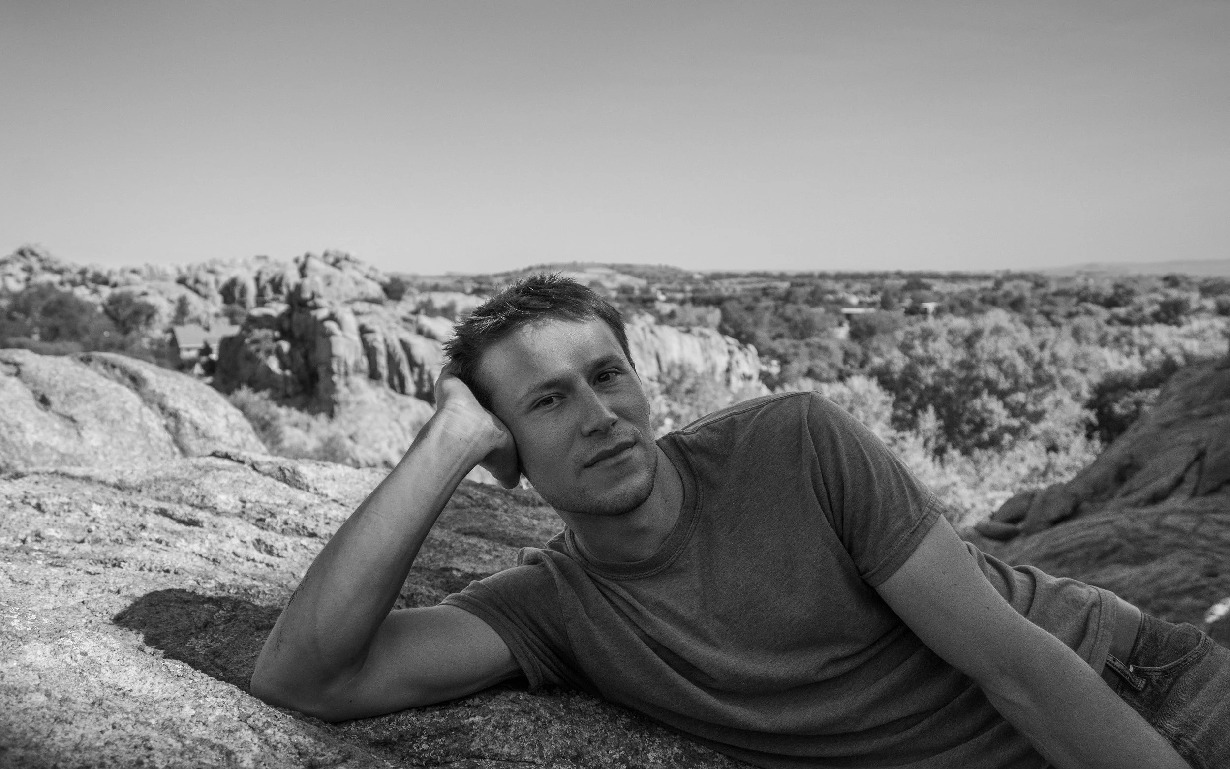 A man lying on a rocky surface in a natural outdoor landscape, resting his head on his hand, with a city or town in the distance.