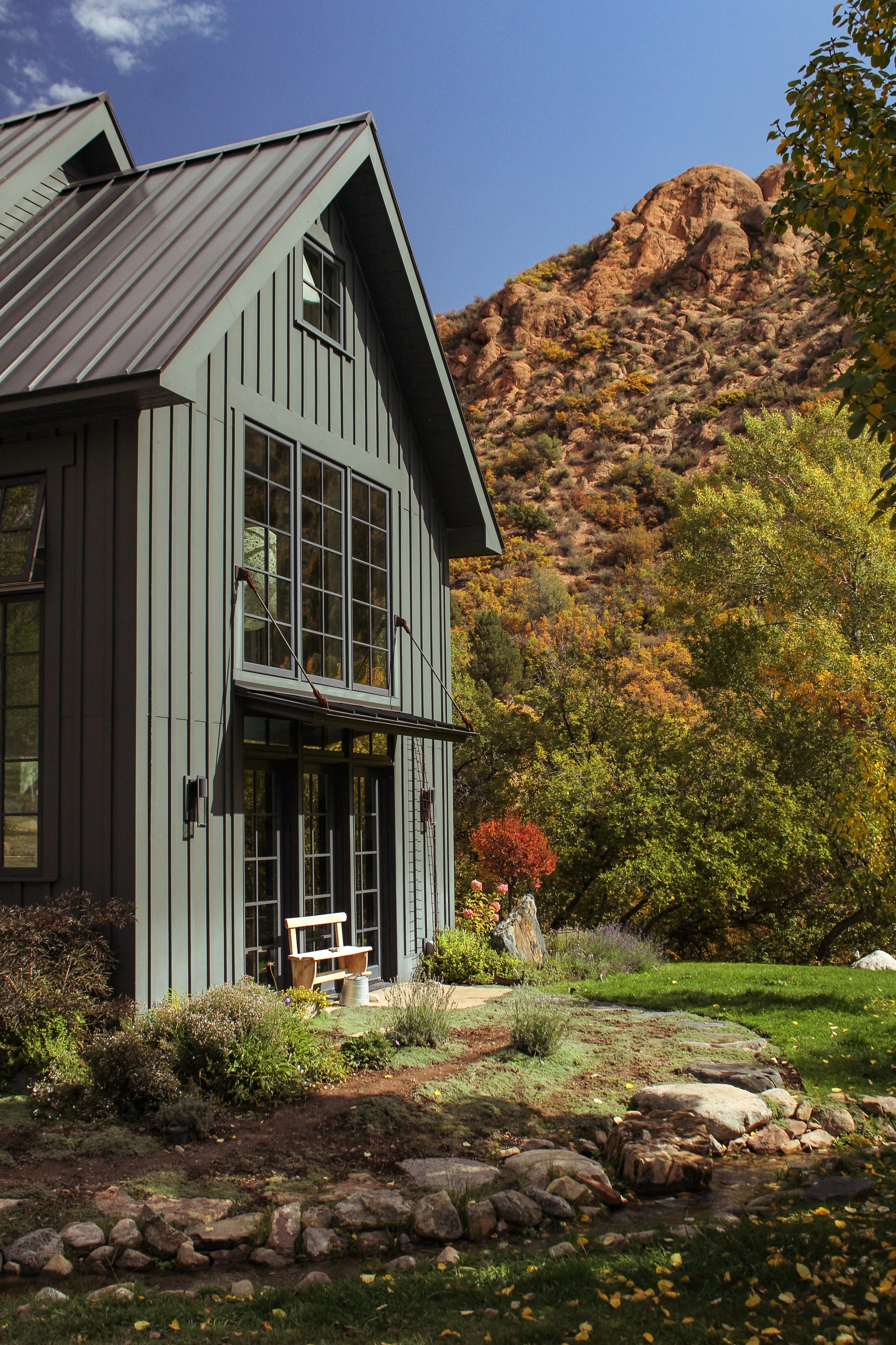 A modern blue house with large windows, situated in a mountainous area during autumn, surrounded by trees with fall foliage and a small stream.