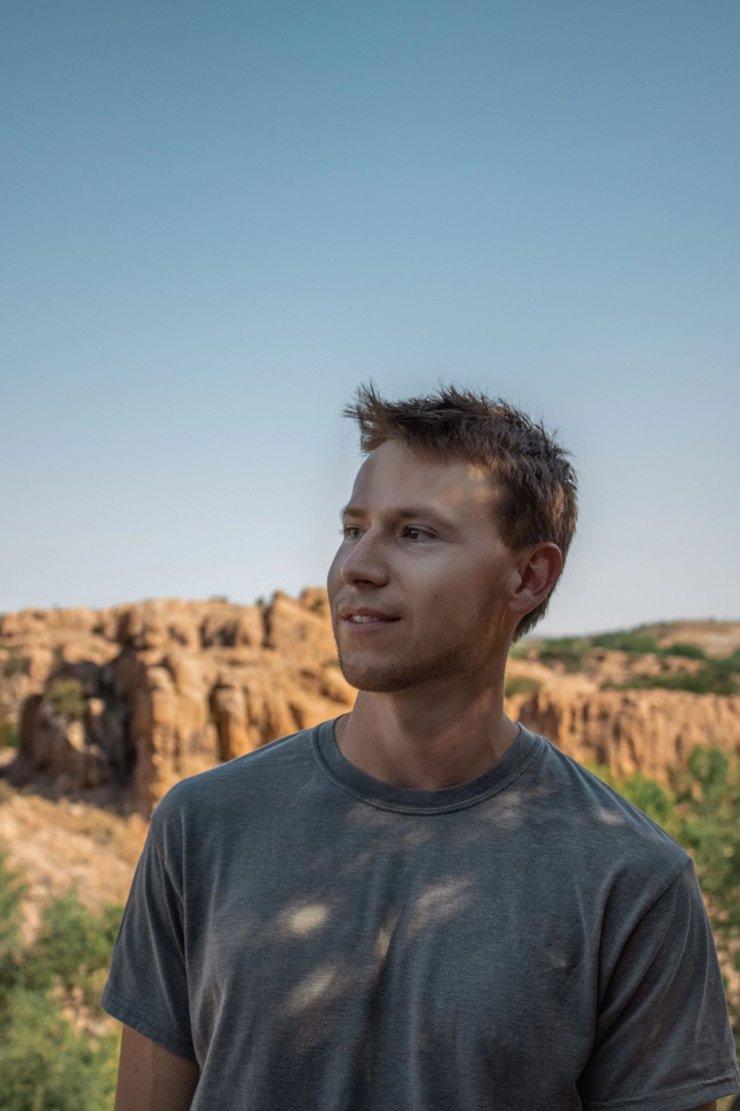 A young man with short, spiky brown hair, wearing a dark grey t-shirt, standing outdoors in a desert landscape with rocky formations and sparse vegetation under a clear blue sky.