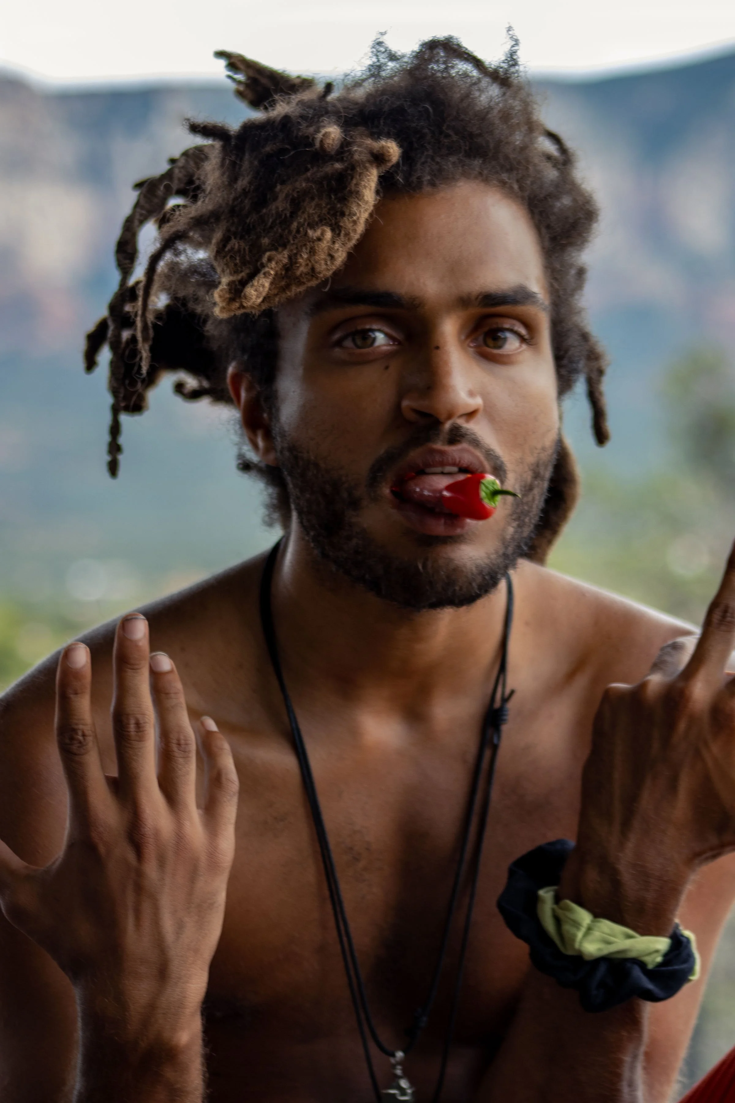 A shirtless man with dreadlocks holding a red chili pepper in his mouth and making hand gestures.