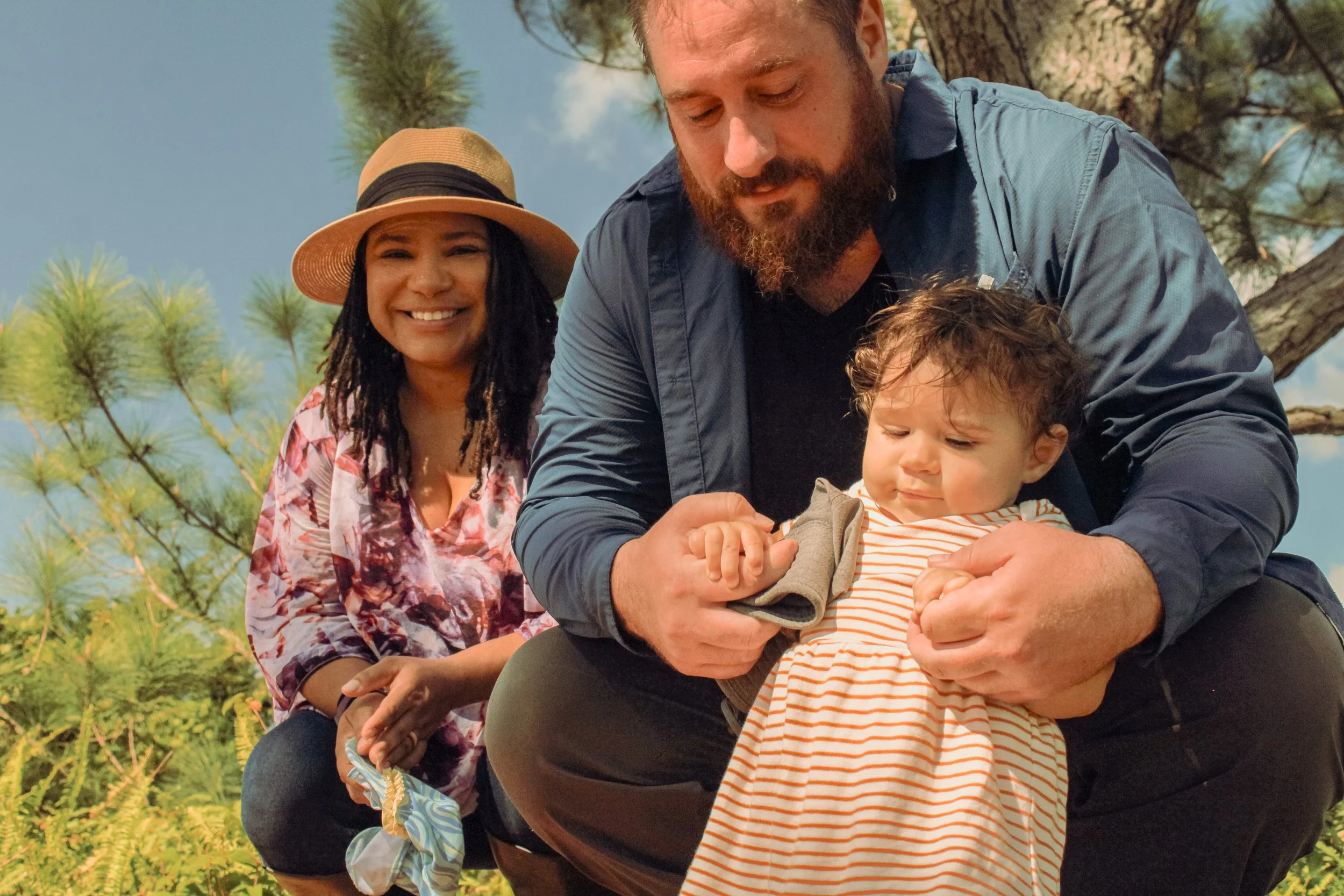 A family outdoors, with a woman in a straw hat and floral shirt smiling, a man holding a young child, and a tree in the background.