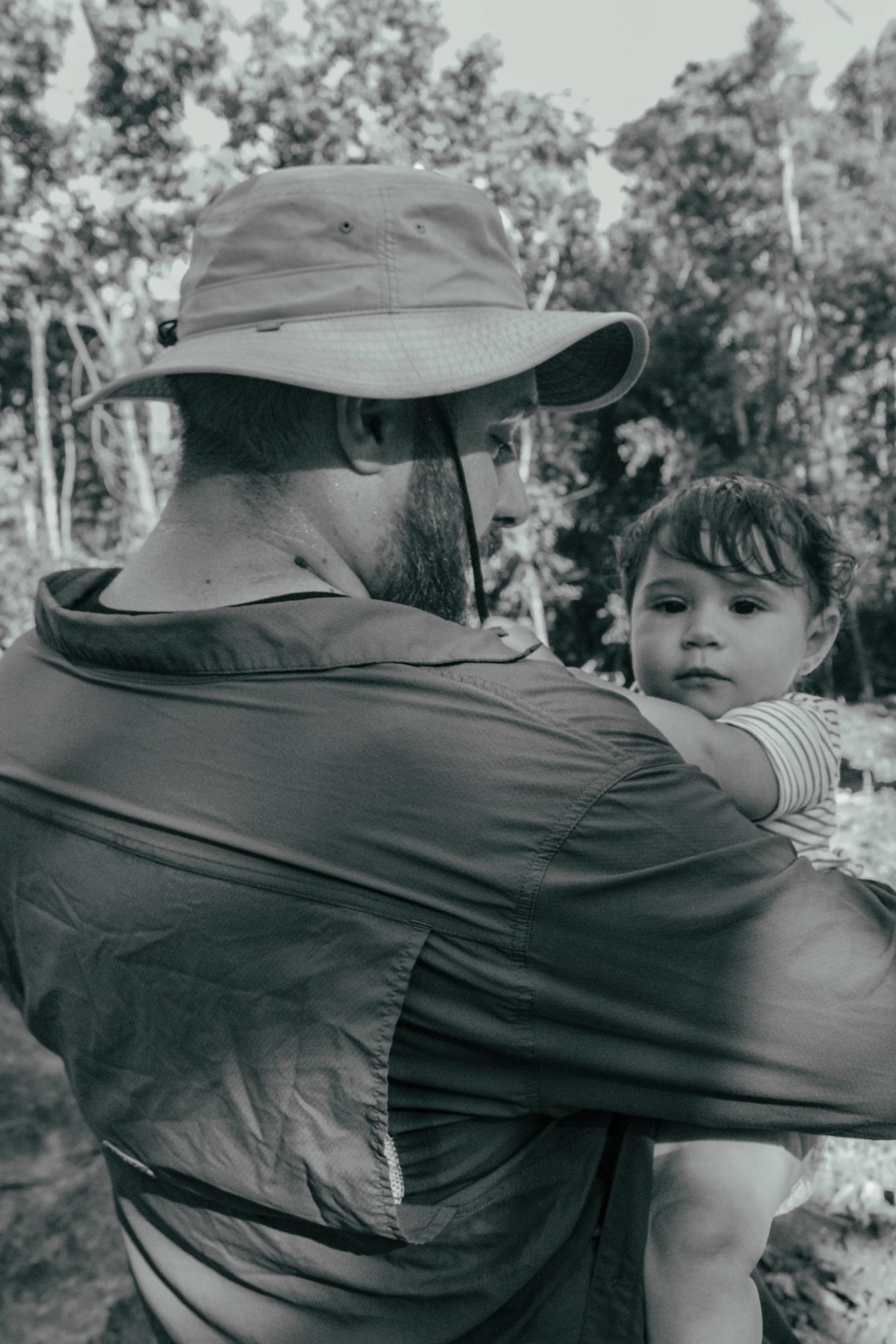 A man with a beard wearing a wide-brimmed hat and outdoor jacket holding a young child in a forested area.