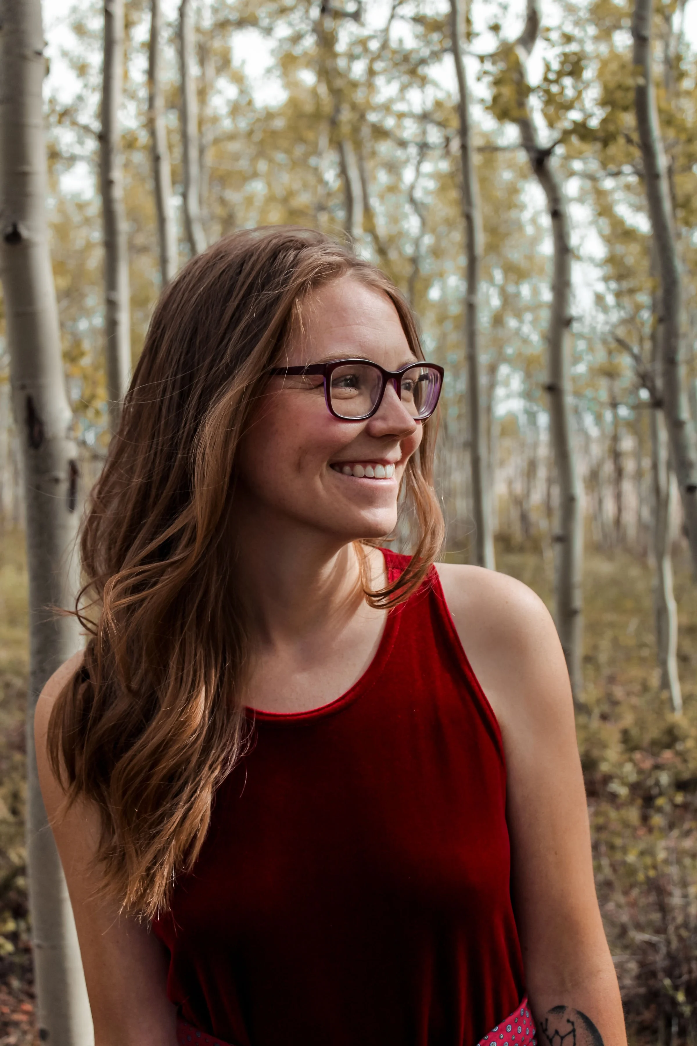 A smiling woman with long red hair, glasses, and a red sleeveless top standing in a forest with tall, light-colored trees and autumn leaves.