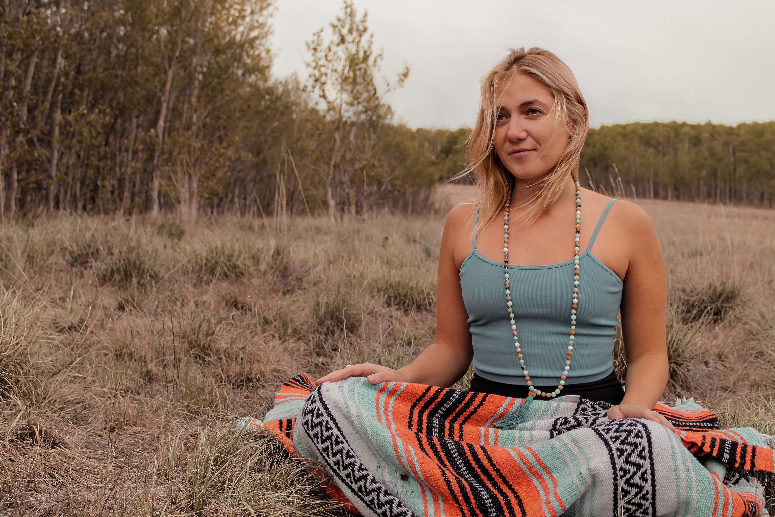 A young woman with blonde hair sitting outdoors on a patterned blanket in a grassy field with trees in the background, wearing a blue tank top and a beaded necklace.