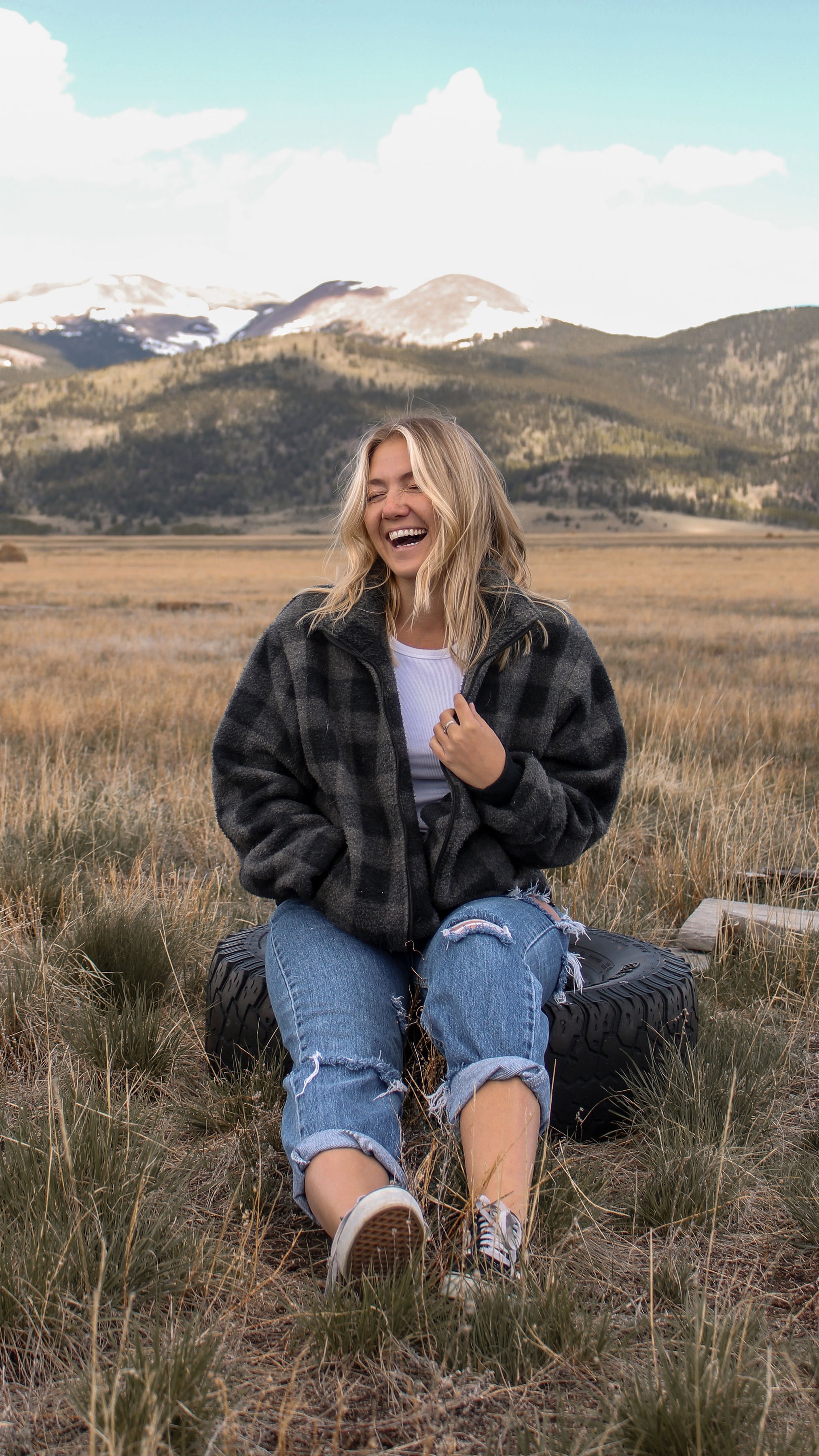 A woman sitting on a tire in an open grassland with mountains in the background, laughing and enjoying the outdoors.