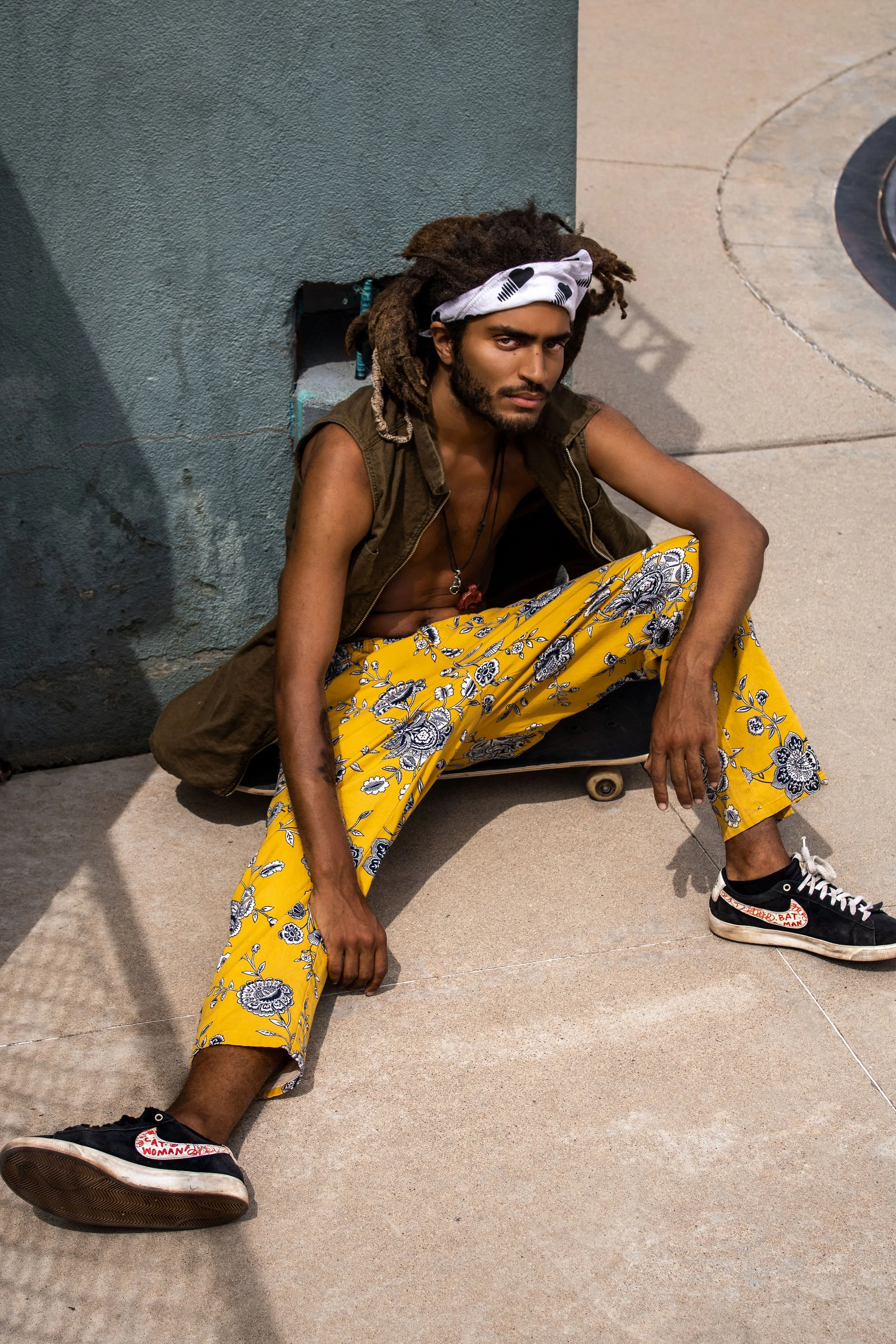 A young man with dreadlocks, a white bandana, and a brown vest sits against a blue wall on a skateboard with his legs spread out, looking at the camera with a serious expression.