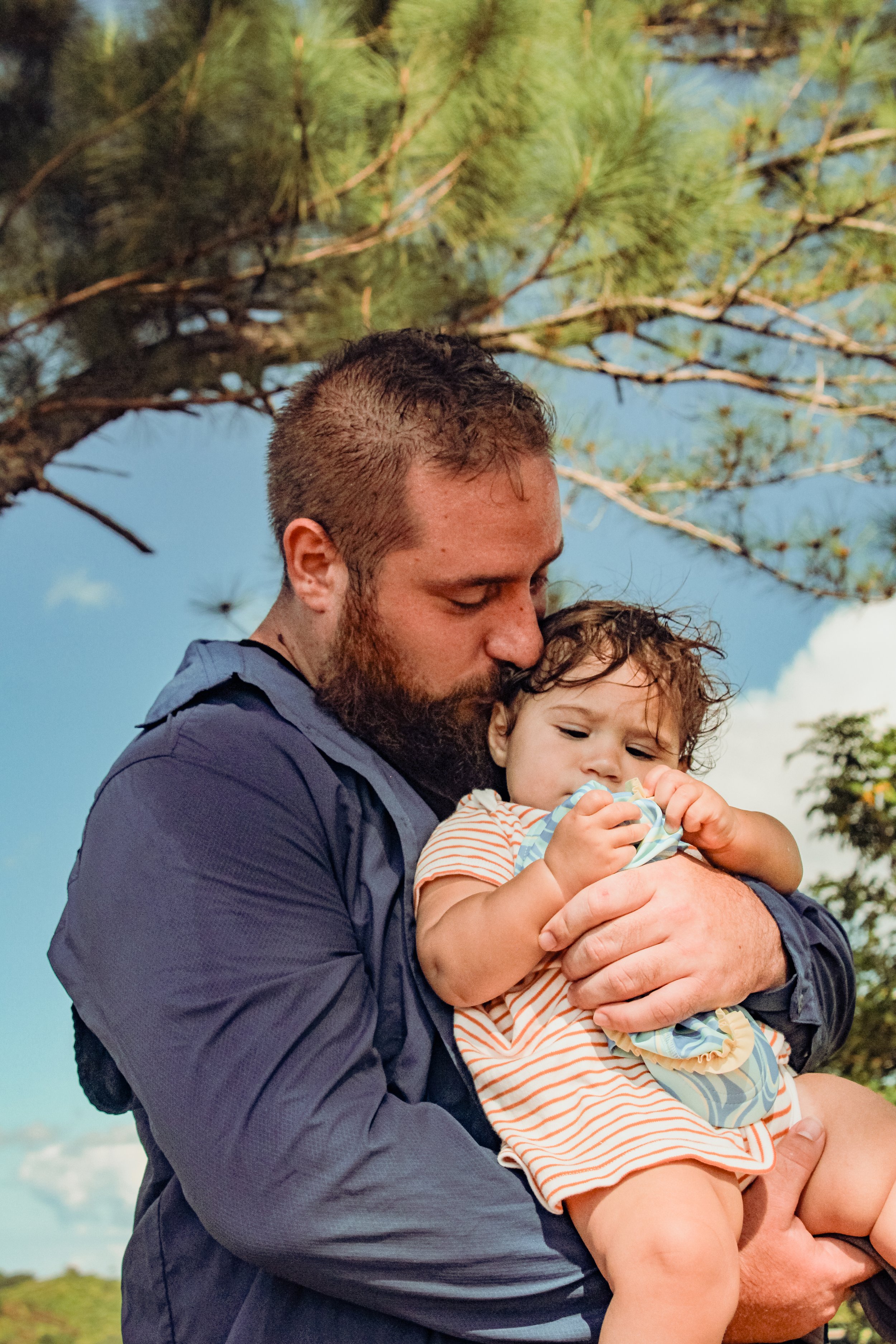A man with a beard holding a young child outdoors near a tree, with a blue sky and some clouds in the background.