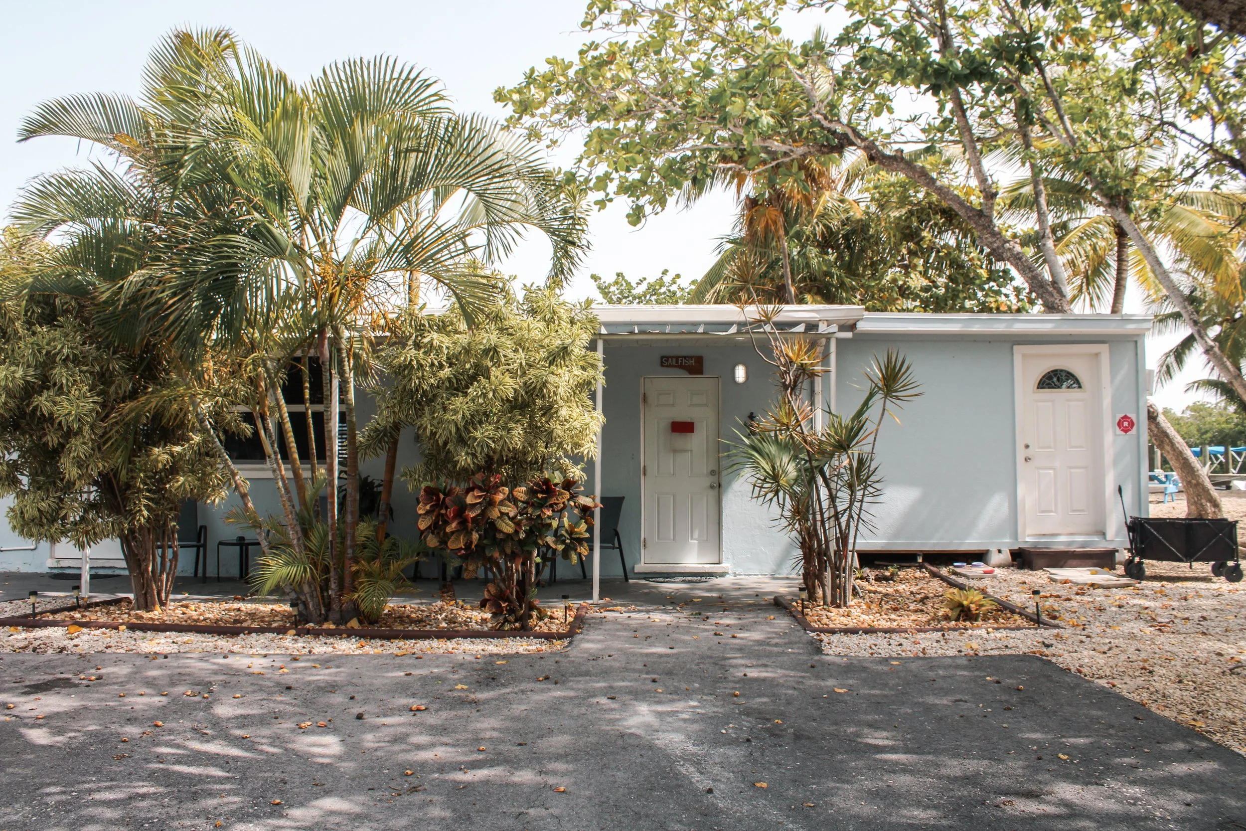 Small, light blue building on a sunny day, surrounded by tropical trees and plants, with a paved driveway and a nearby beach area.