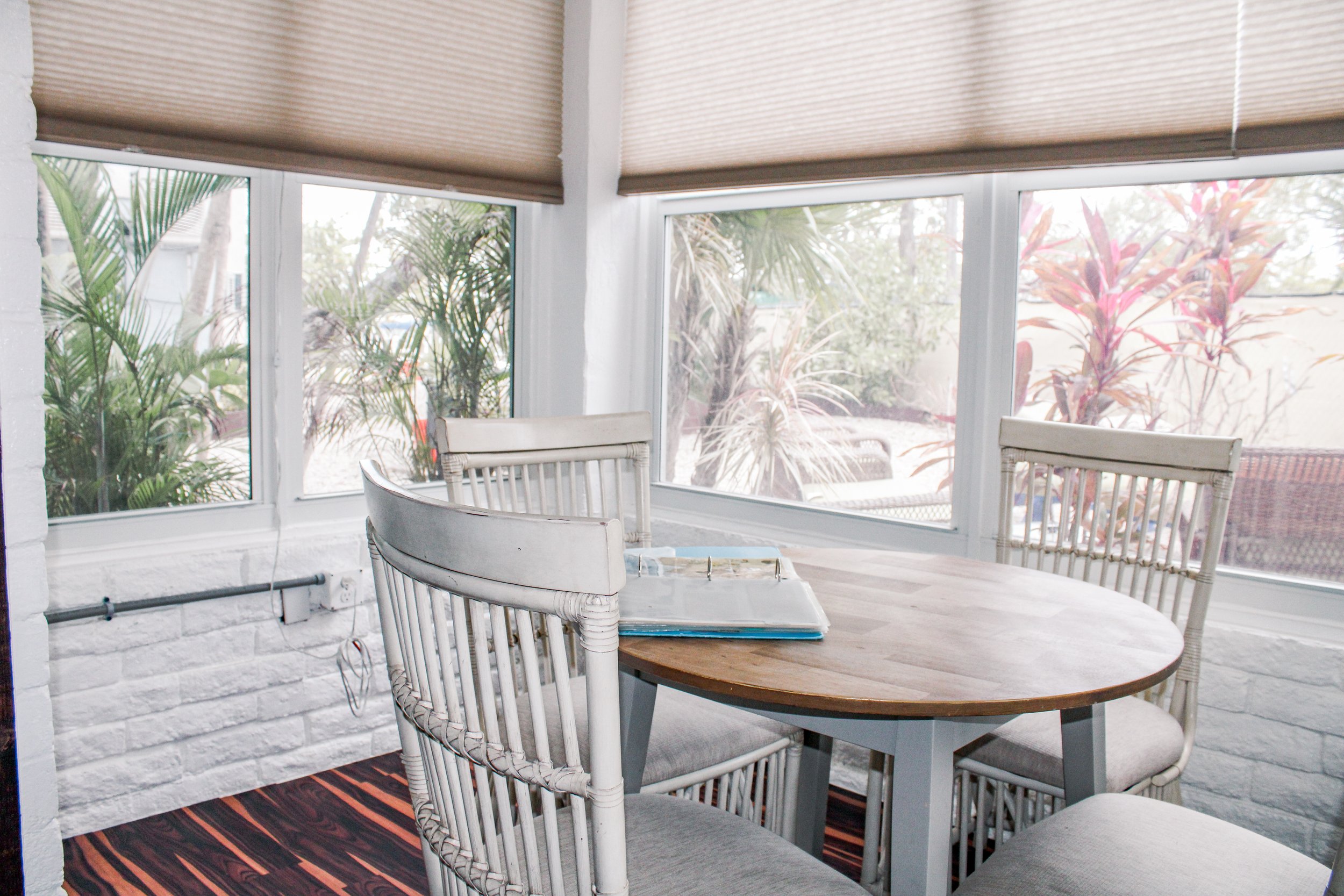 Dining area with a round wooden table, four white chairs with cushions, large windows with beige blinds, view of lush plants outside, and a wooden floor.