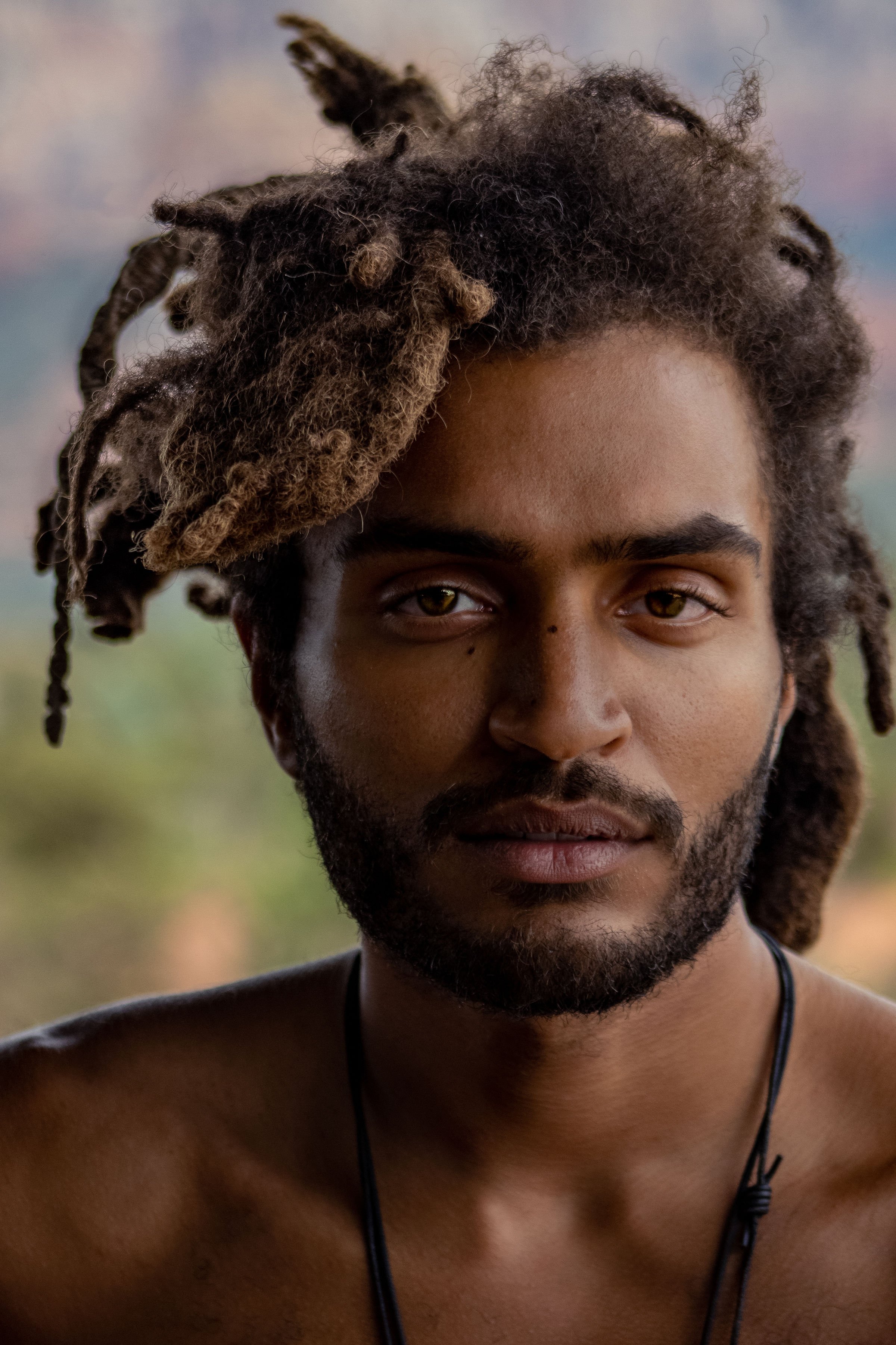Close-up portrait of a young man with dreadlocks and a beard, looking directly at the camera, with a blurred outdoor background.