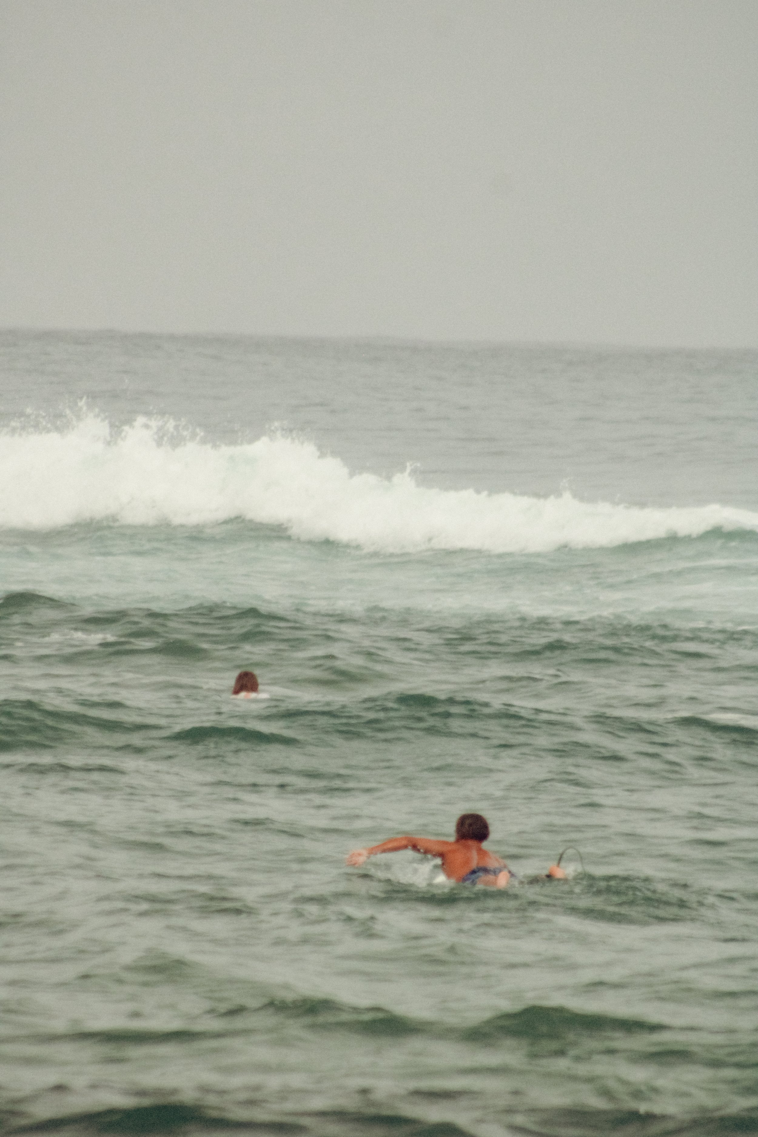 Two surfers swimming in the ocean with waves in the background.