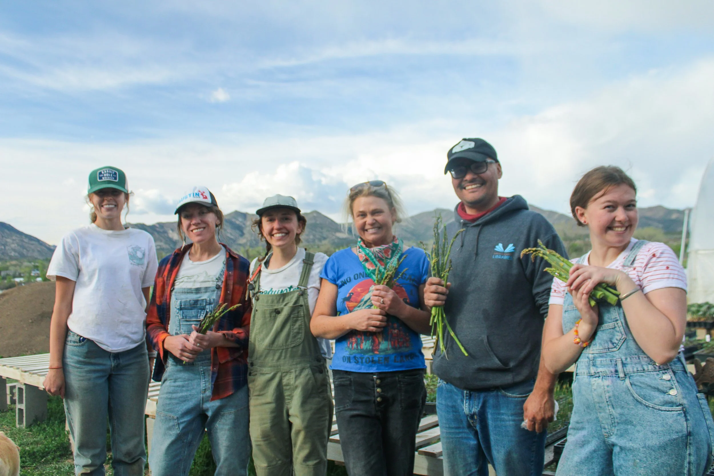 Group of six people standing outdoors, smiling, holding plants, with mountains and blue sky in the background.