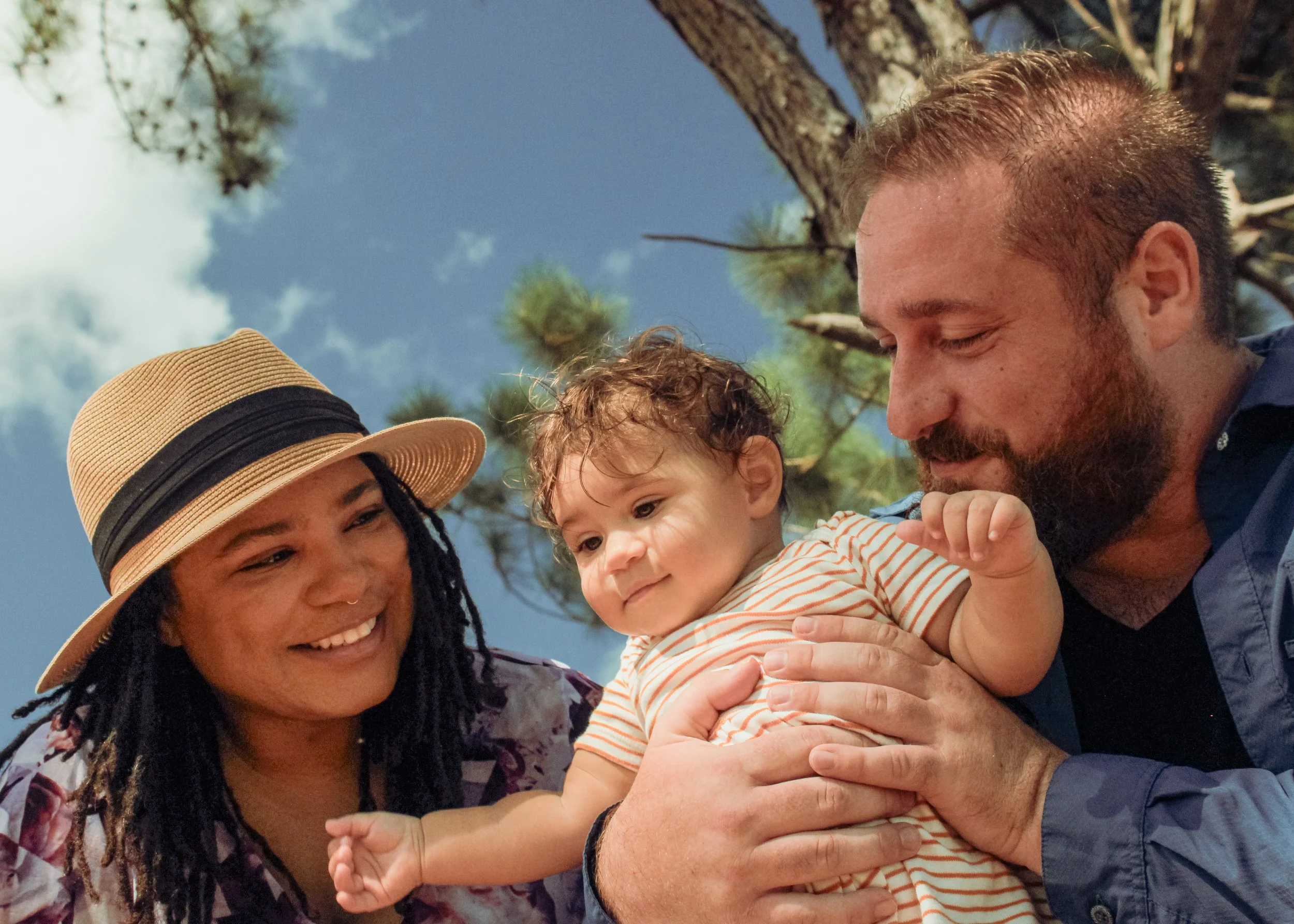 A family outdoors, with a woman wearing a straw hat, a man holding a young child, and trees in the background.