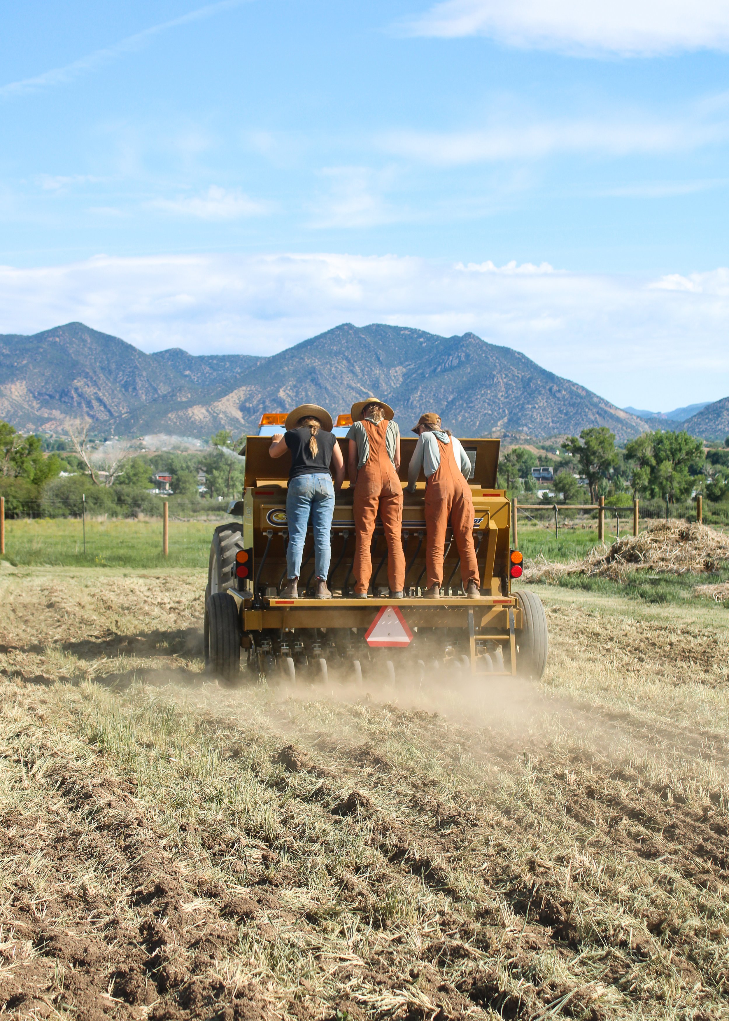 Three people in overalls and hats working on a tractor in a field with mountains in the background.