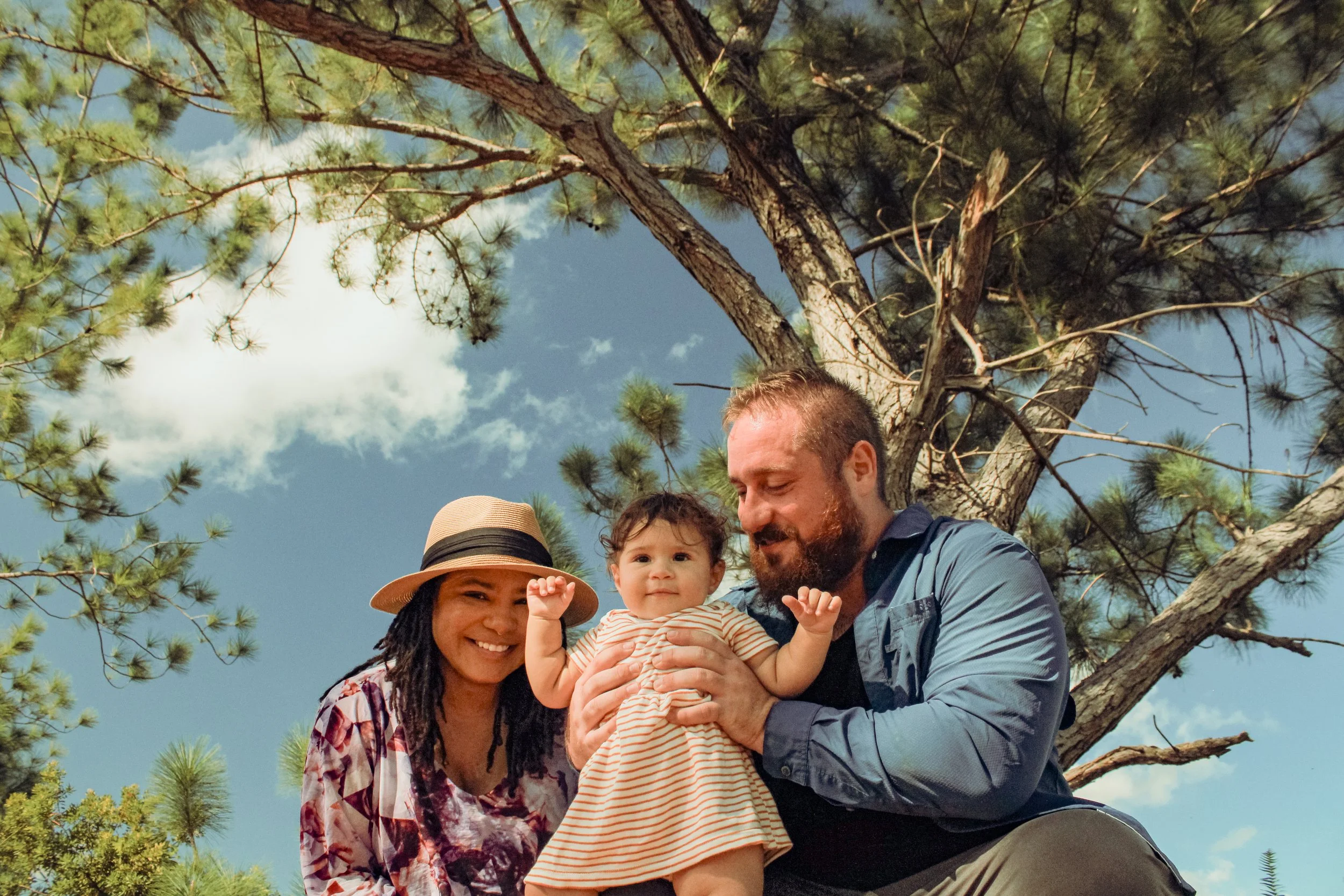 A family of three outdoors with a tall pine tree and blue sky in the background. A woman with curly hair and a hat smiles, a man with a beard holds a young girl with dark hair, all appearing happy.