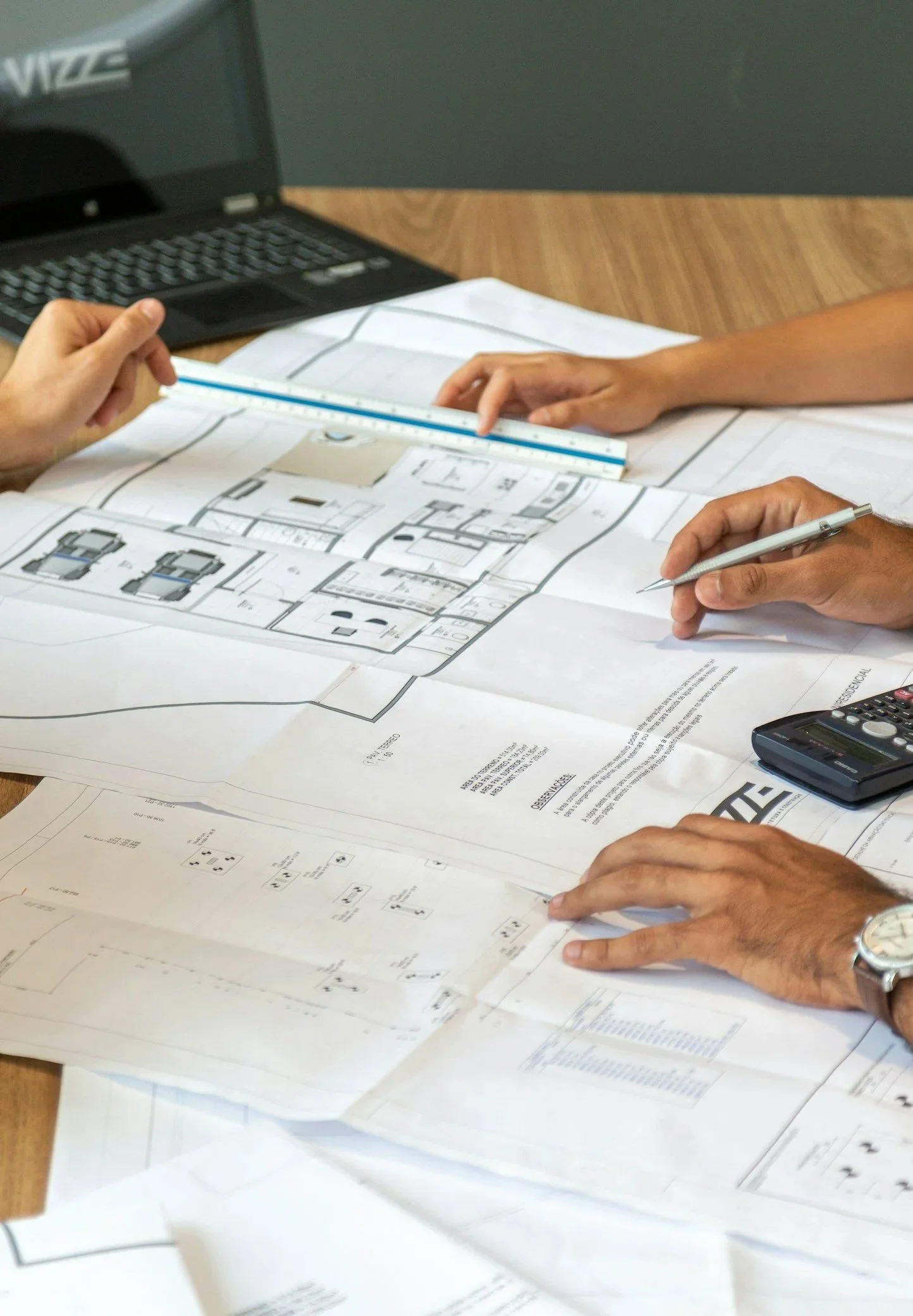 Two people reviewing architectural blueprints on a table, with a calculator, pen, and computer nearby.