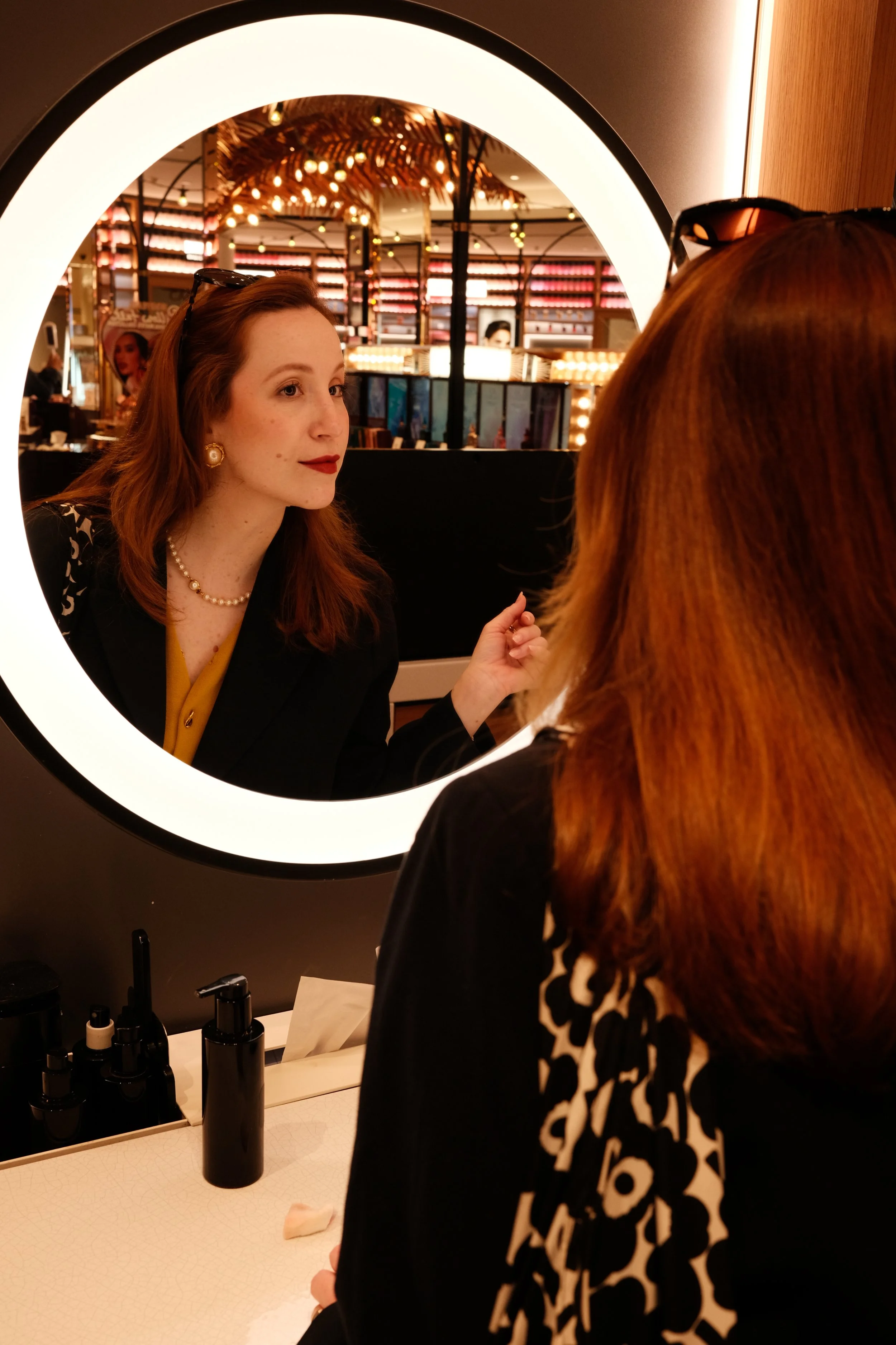 Mulher com cabelo castanho claro e roupas elegantes olhando no espelho circular com iluminação, com um ambiente decorado com luzes e plantas ao fundo.