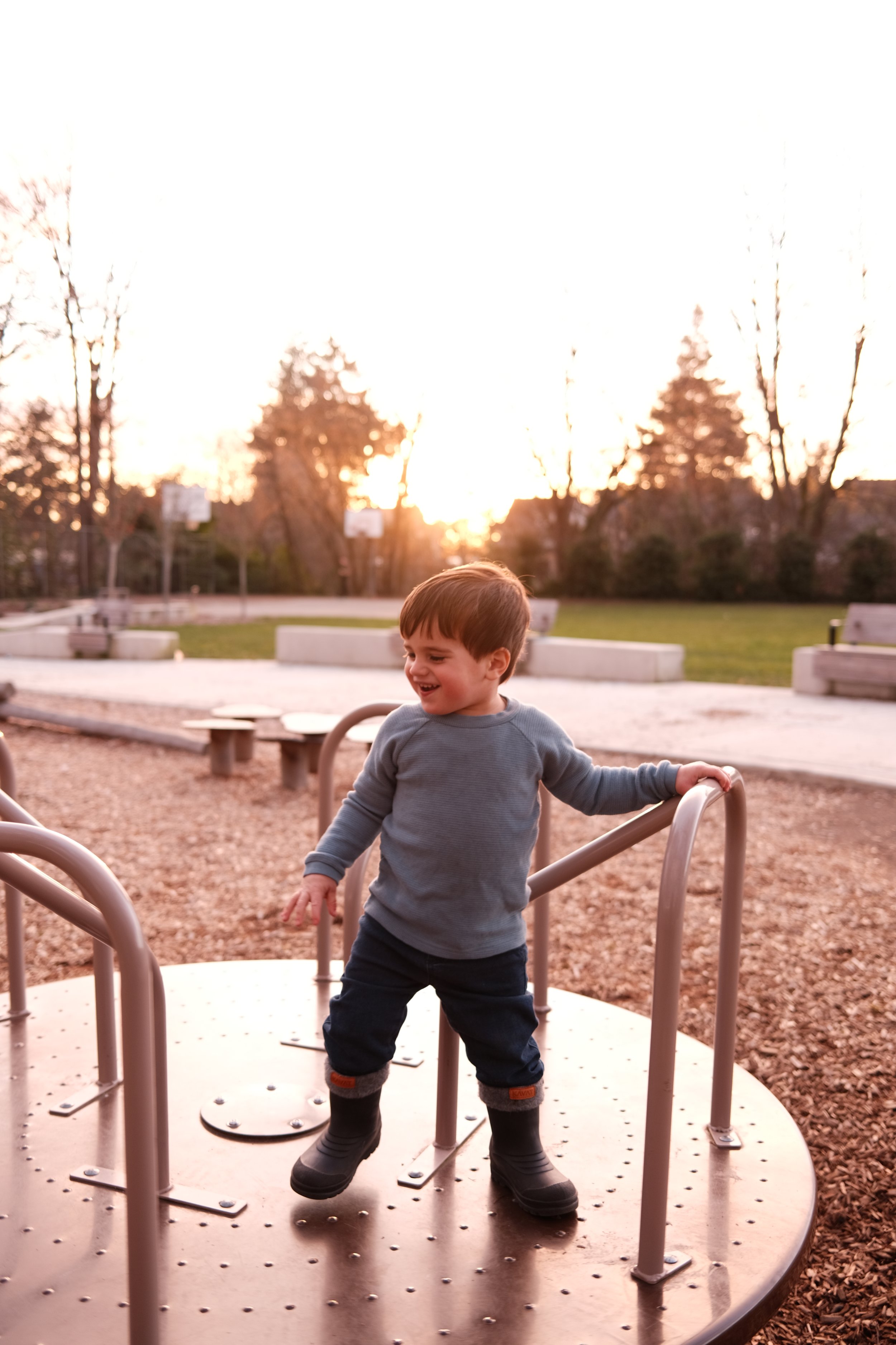 Menino brincando em um parque ao entardecer, sorrindo e segurando uma grade de brinquedo.
