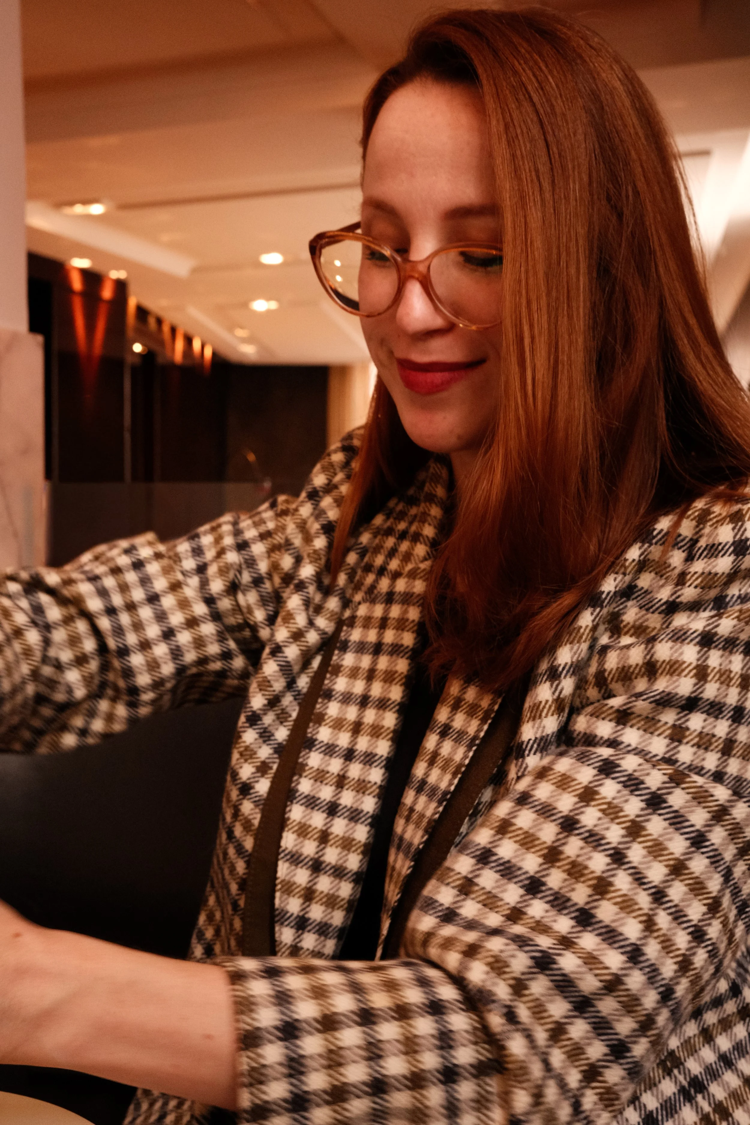 Mulher de cabelo castanho-avermelhado, usando óculos de armação tortoise eblusada com padrão xadrez em tons de marrom, bege e preto, sorrindo ligeiramente enquanto interage com algo fora da câmera em um ambiente interno com iluminação quente.