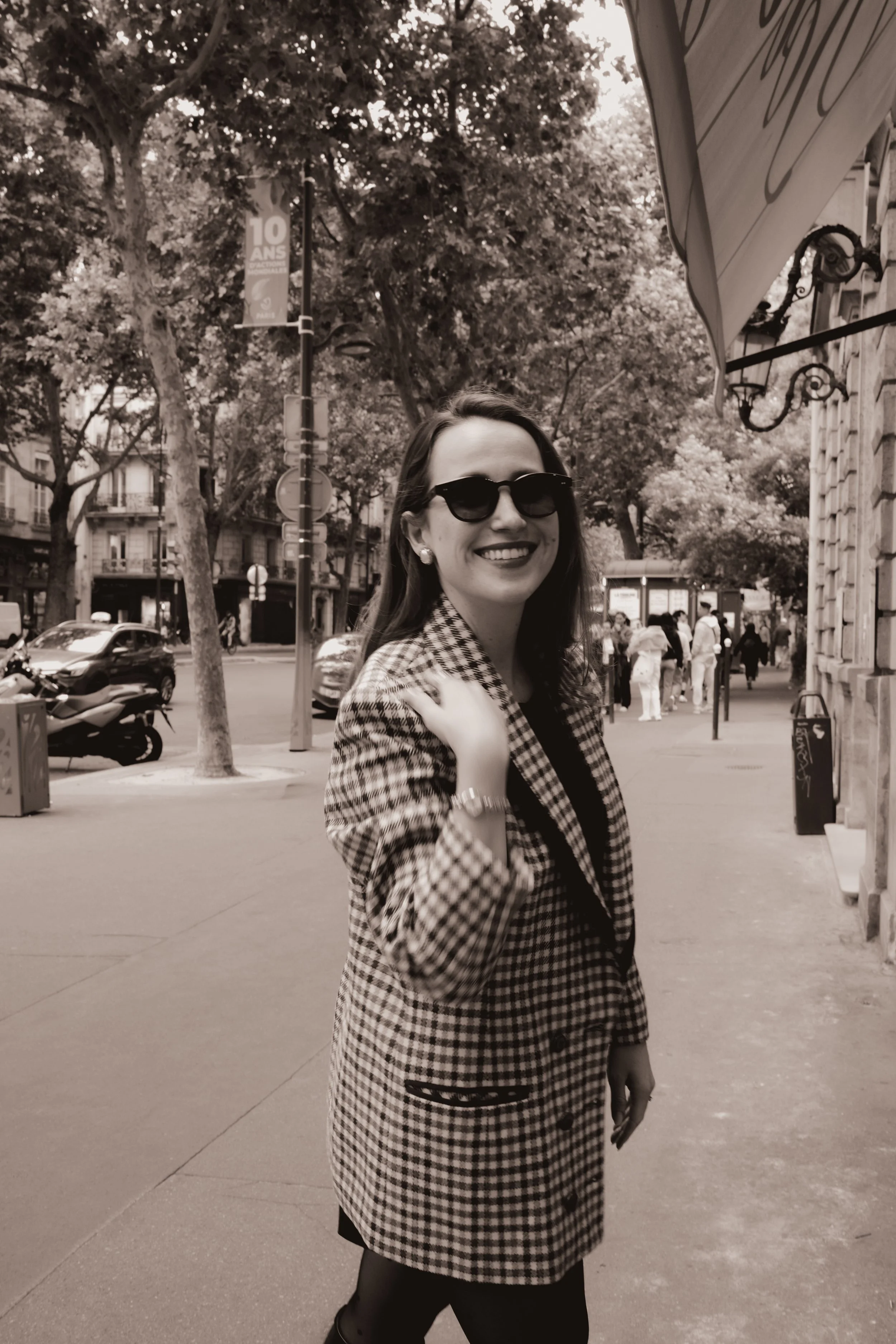 Mulher sorridente com óculos escuros, casaco xadrez, na calçada de uma rua arborizada, com pessoas ao fundo e carros estacionados.