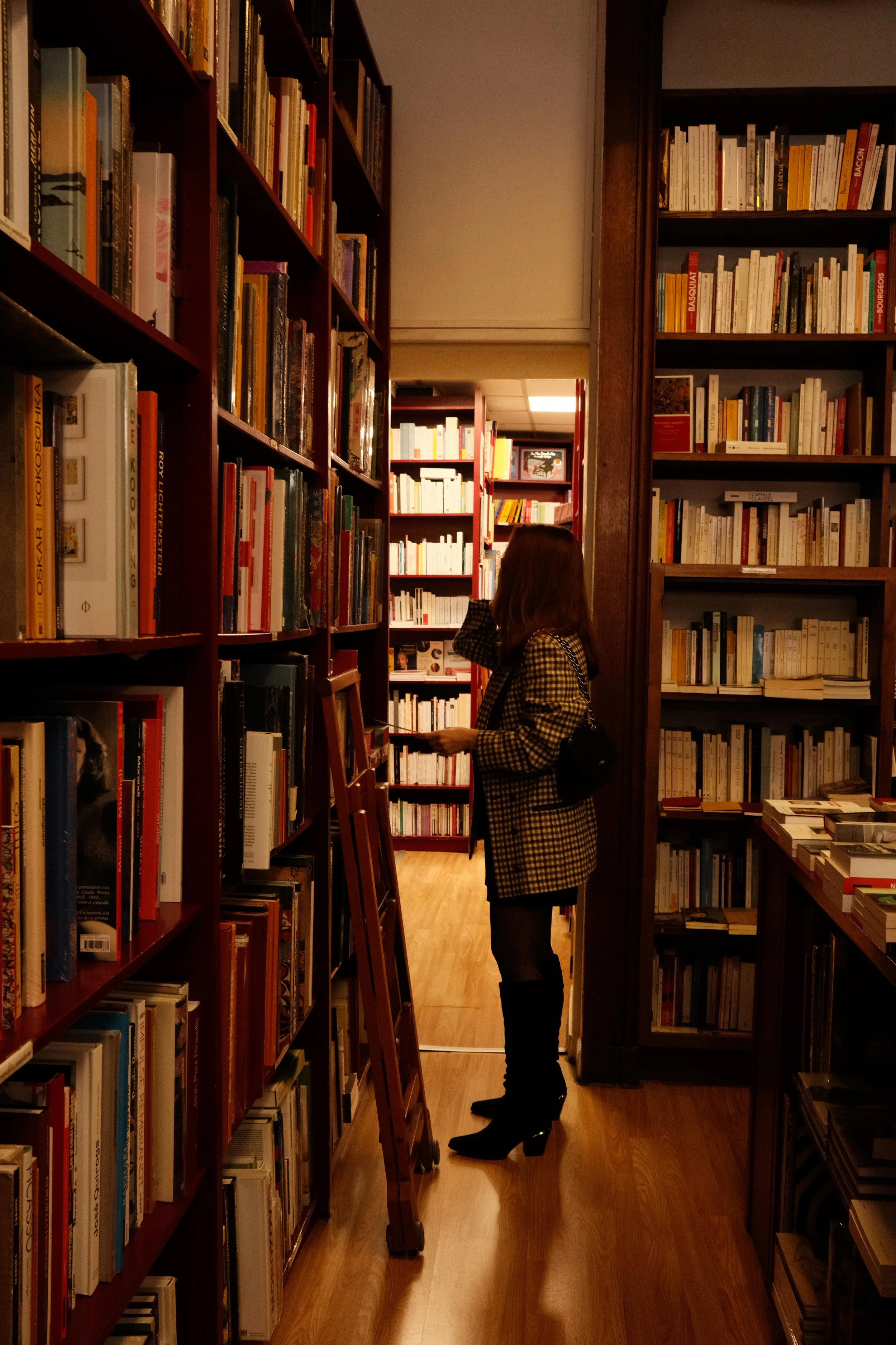 Pessoa feminina de meia-idade, com cabelo castanho, vestindo jaqueta xadrez em tom amarelado, preto e botas de salto, segurando um livro, em uma biblioteca com prateleiras repletas de livros ao redor.