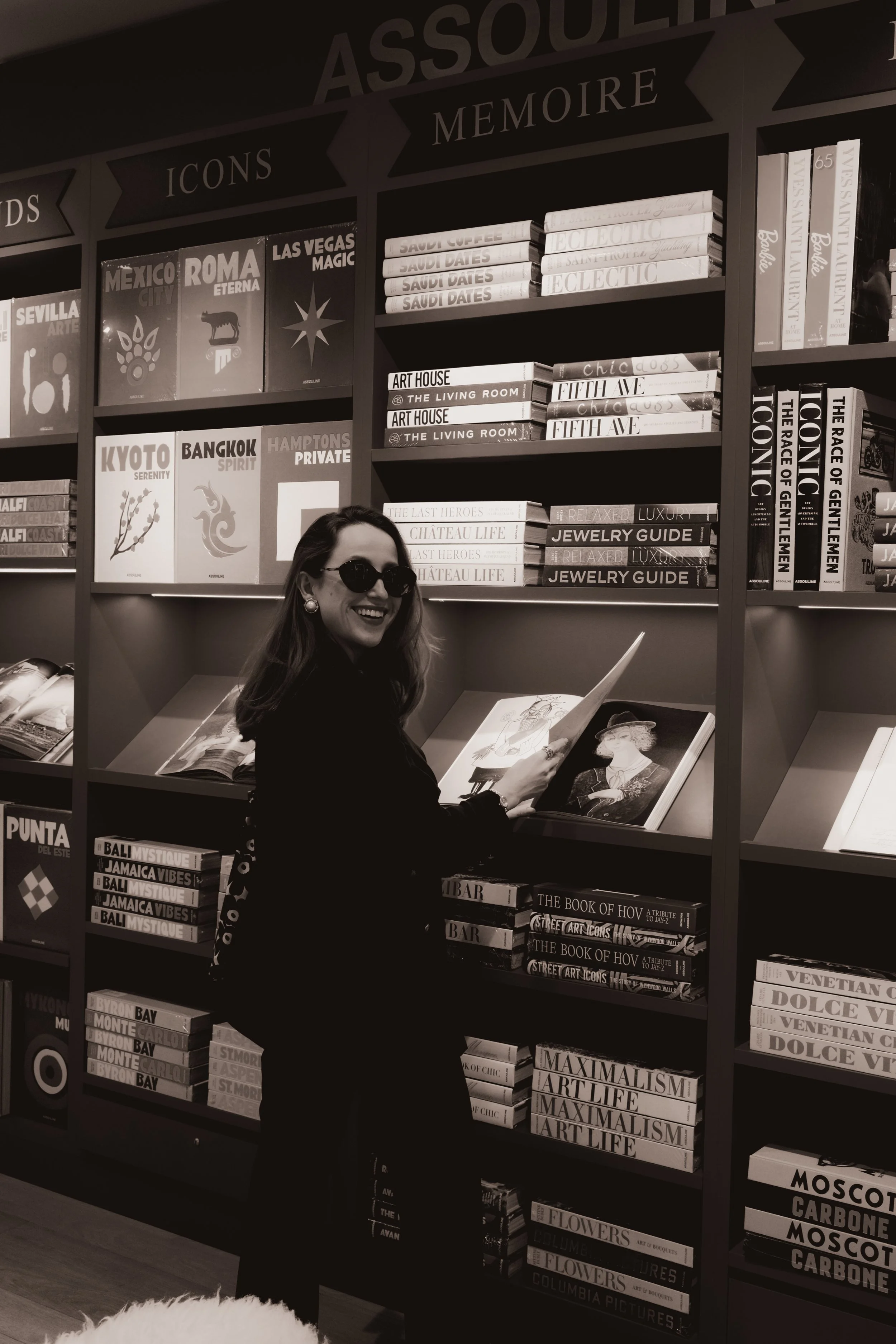 Mulher sorridente de óculos escuros, de cabelo longo, vestindo roupas escuras, segurando um livro aberto na frente de uma estante de livros em uma livraria.