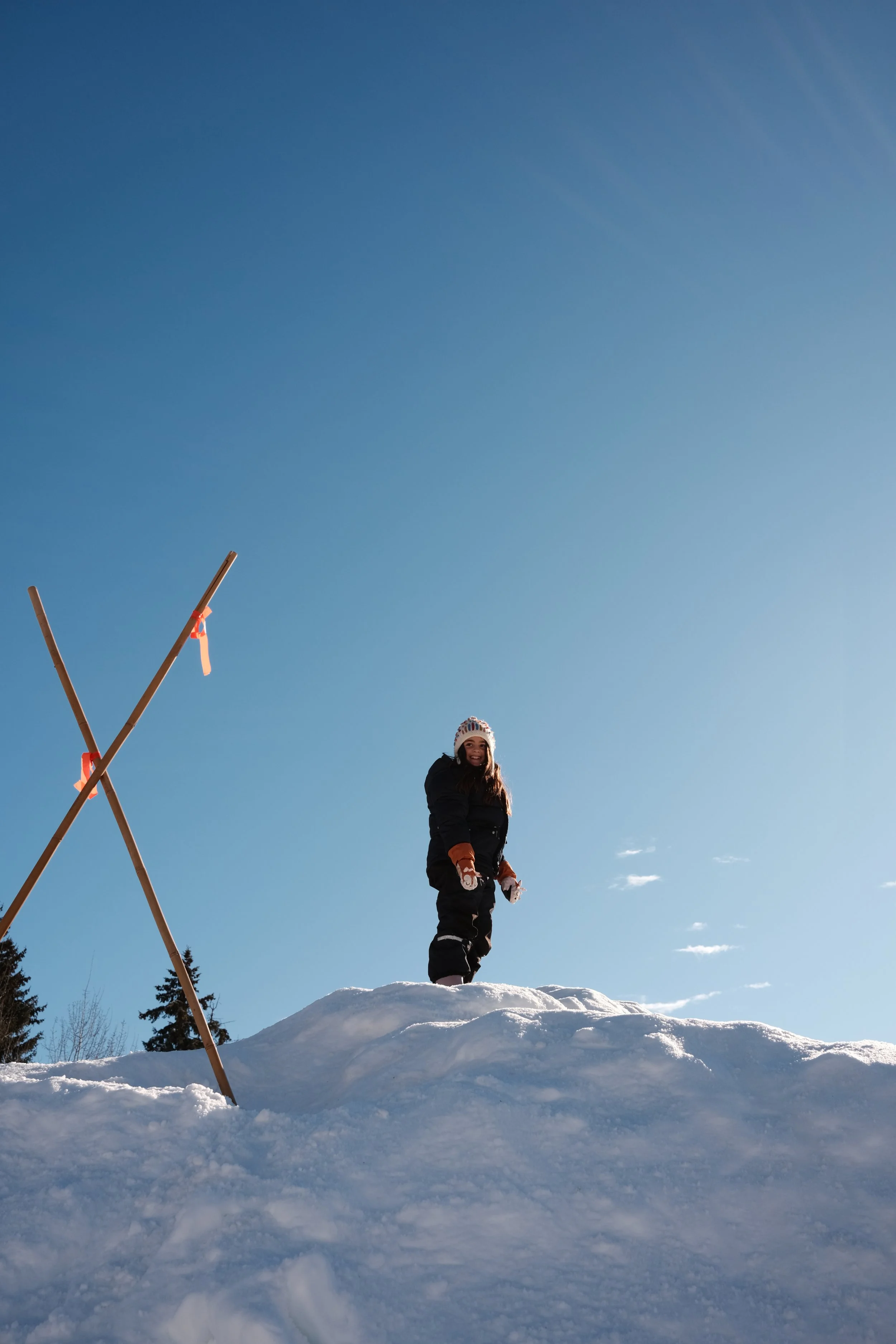 Pessoa feliz vestida com roupas de inverno de pé na neve sob um céu claro, ao lado de uma estrutura de madeira com fitas coloridas.