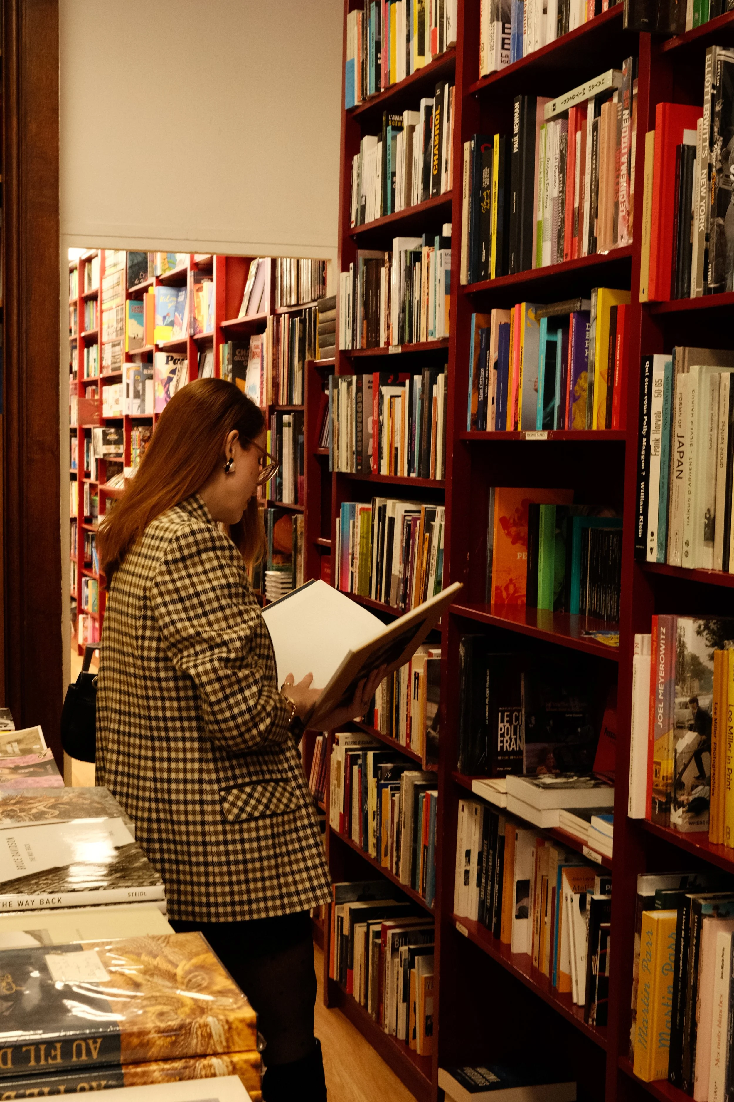 Mulher com cabelo castanho, usando óculos e blazer de xadrez, lendo um livro em uma livraria rodeada por prateleiras de livros.