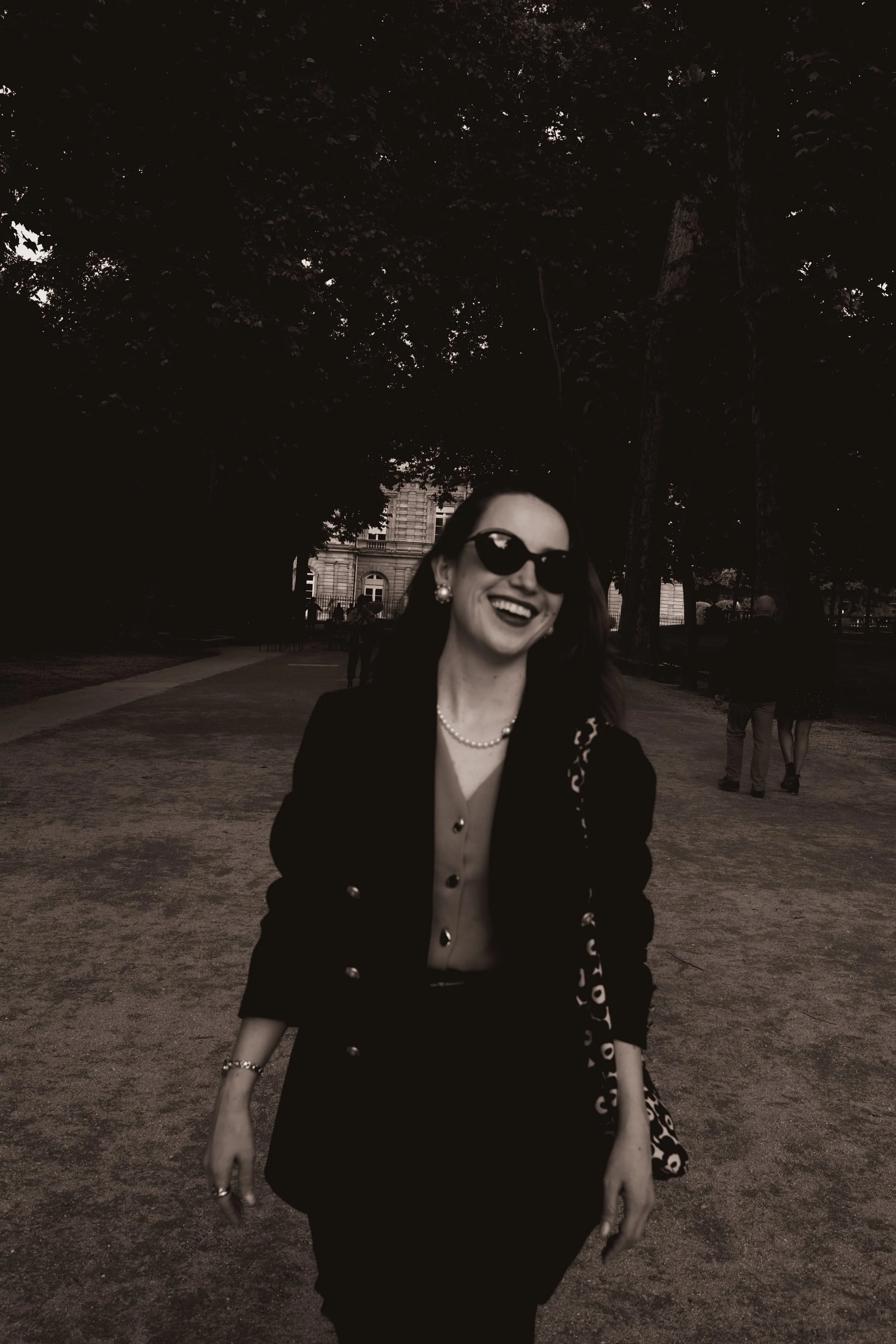 Mulher sorrindo usando óculos escuros, jaqueta preta, saia preta e acessórios na rua com árvores ao fundo.