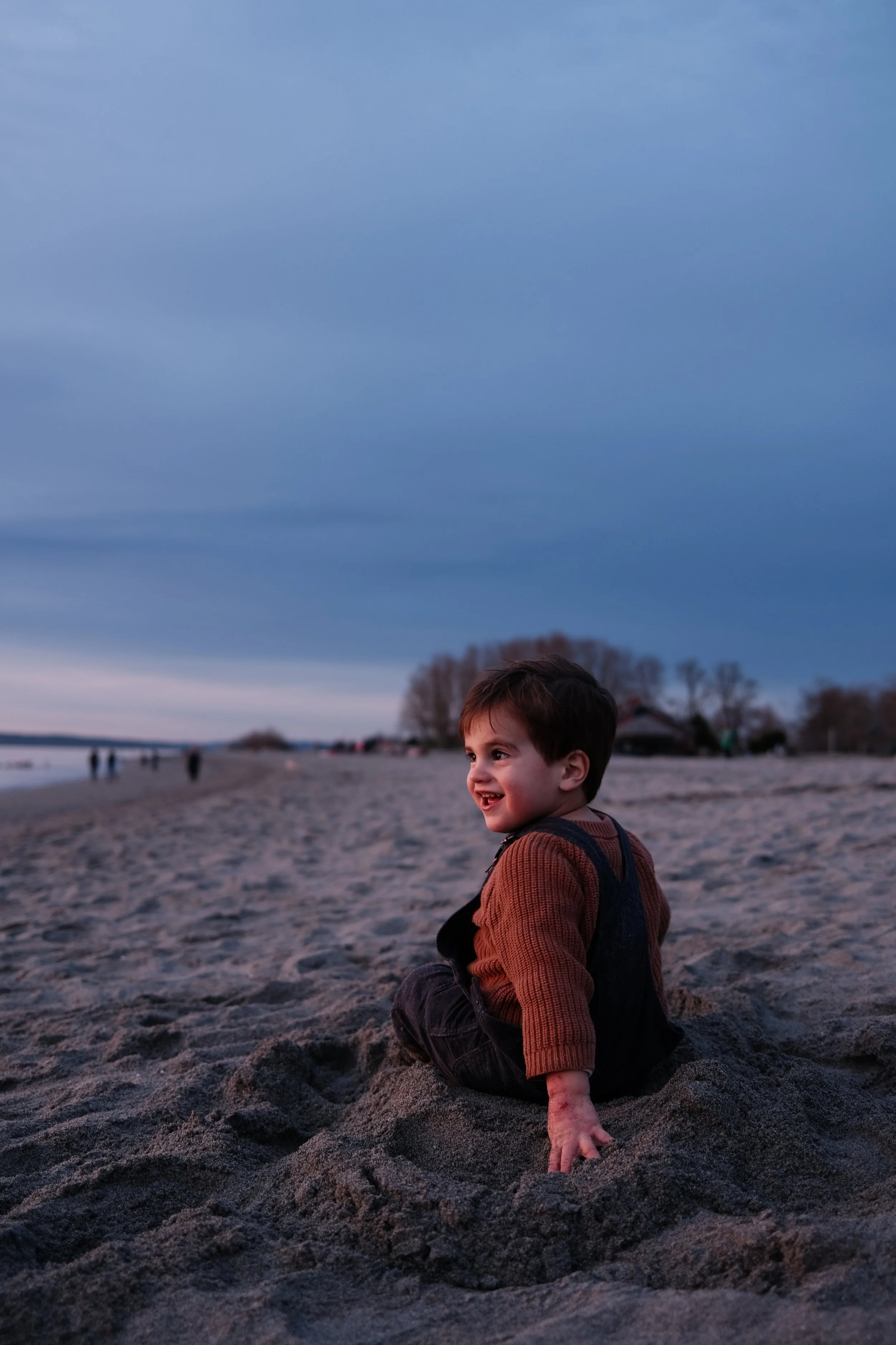 Criança rindo enquanto senta na praia ao entardecer, com céu nublado ao fundo.