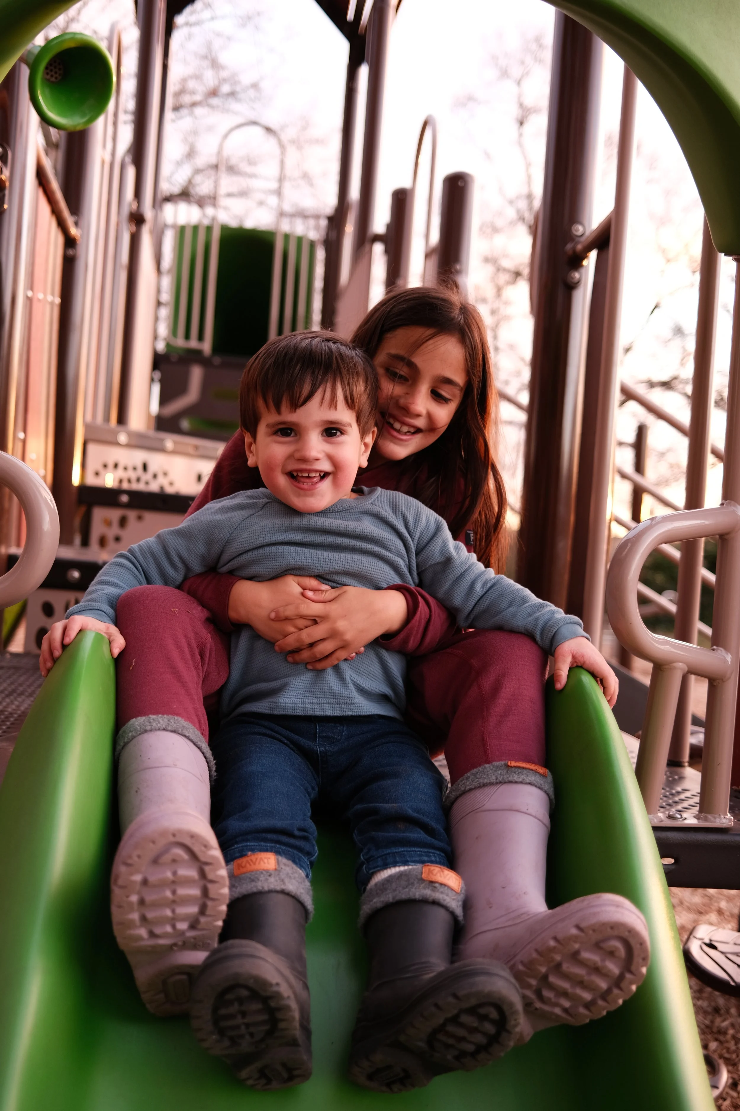 Duas crianças descendo juntas por um escorregador de parque infantil de madeira e metal, sorrindo felizes, com árvores ao fundo.