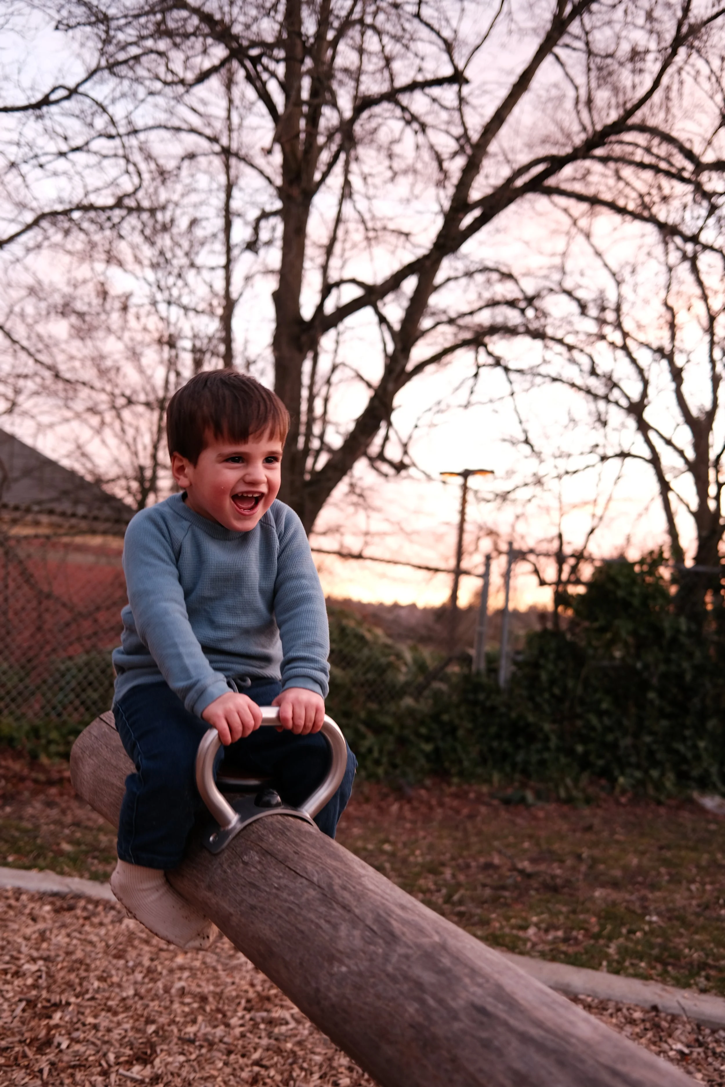 Criança sorridente brincando em parque no final da tarde, sentado em um tronco de árvore, com árvores secas ao fundo e céu com cores de pôr do sol.