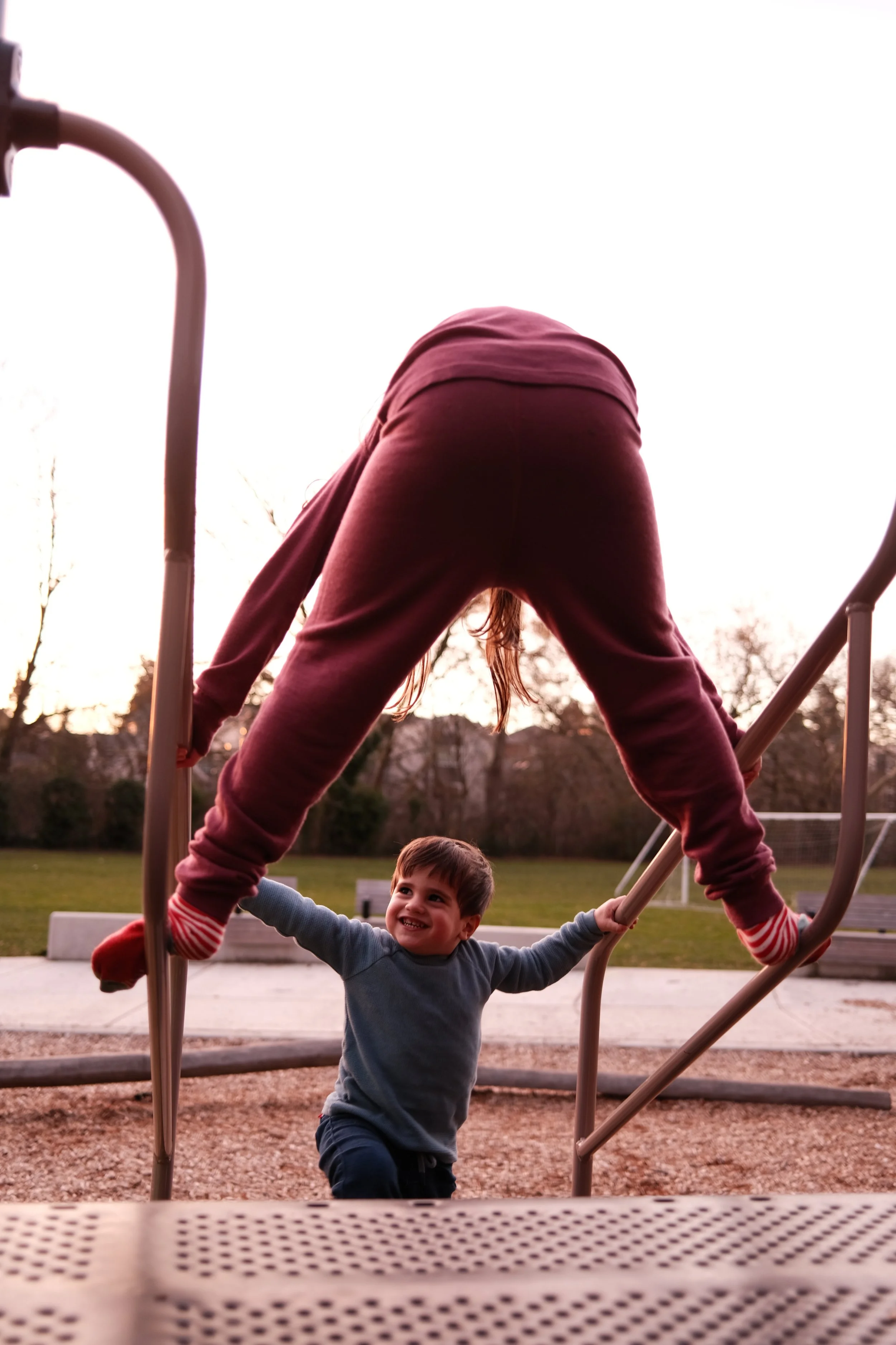 Crianças brincando em parque ao pôr do sol, uma delas se pendurando em uma estrutura de play e a outra sorrindo olhando para ela.