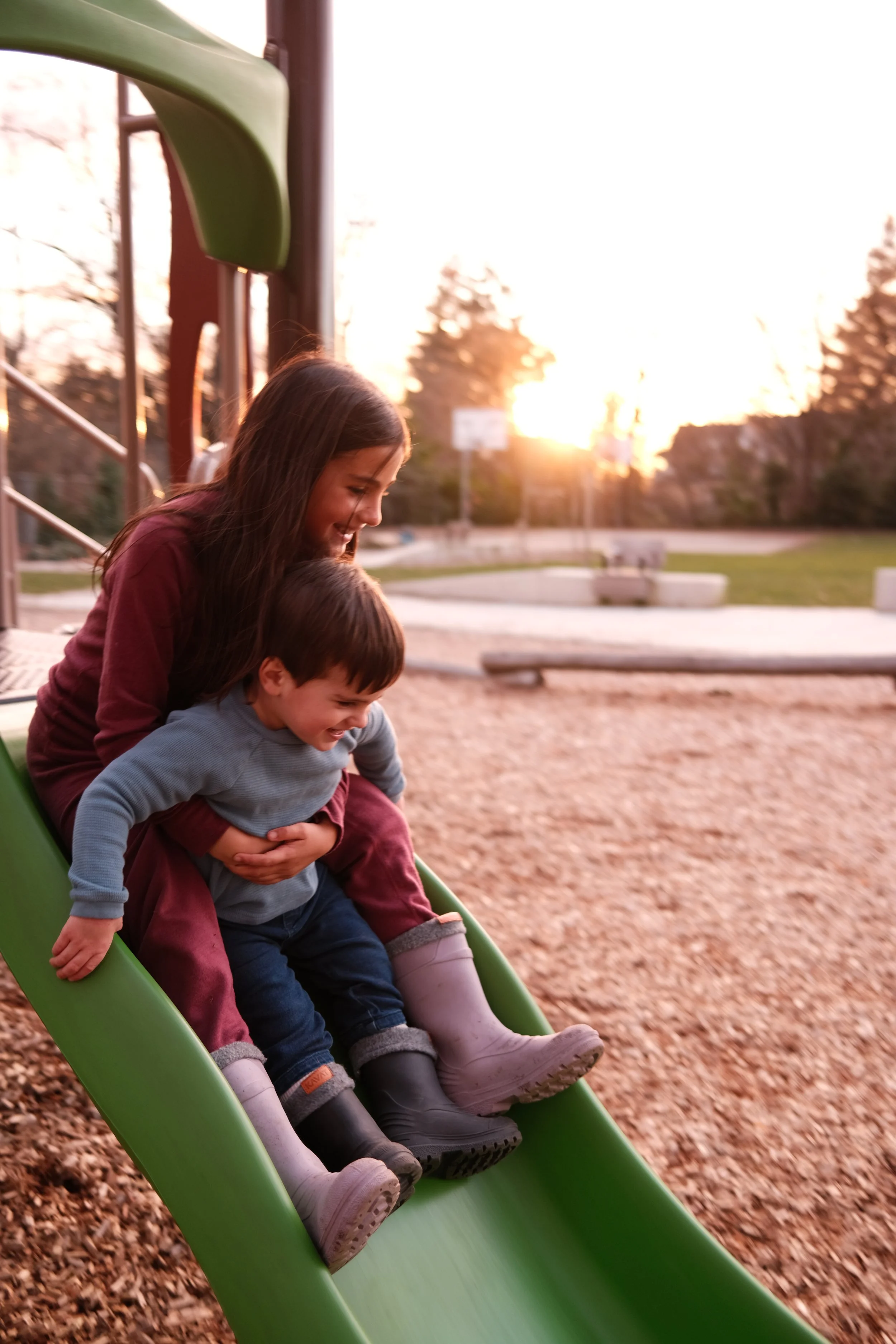 Duas crianças felizes descendo juntas por um escorregador verde em um parque ao pôr do sol.
