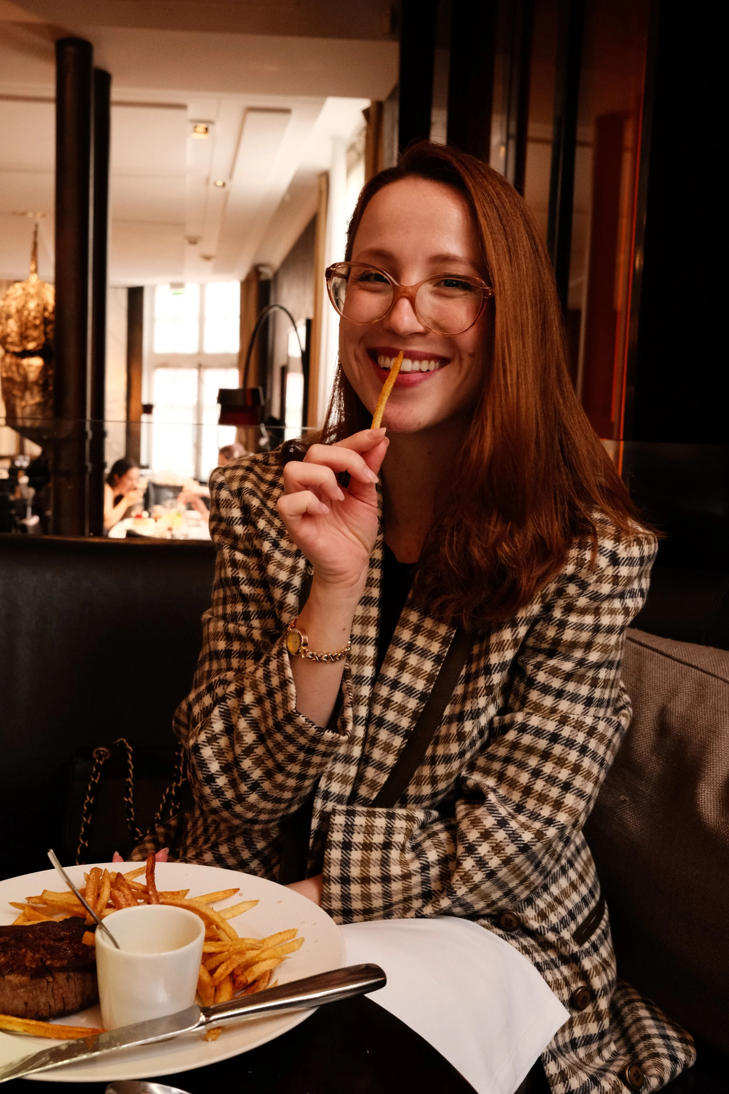 Mulher sorrindo, usando óculos e blazer xadrez, segurando um palito de batata-frita na boca em restaurante, com prato de comida, batatas fritas e carne na mesa.